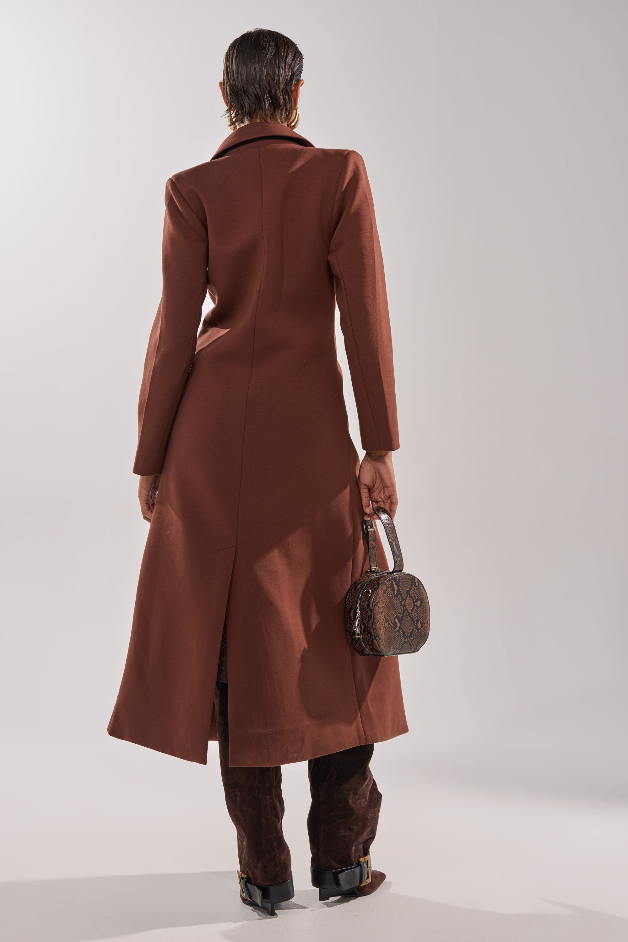A person stands facing away, wearing the COCOA BUTTER SCUBA TRENCH IN BROWN with dark pants and boots, holding a small brown textured handbag in their right hand against a neutral background. 4