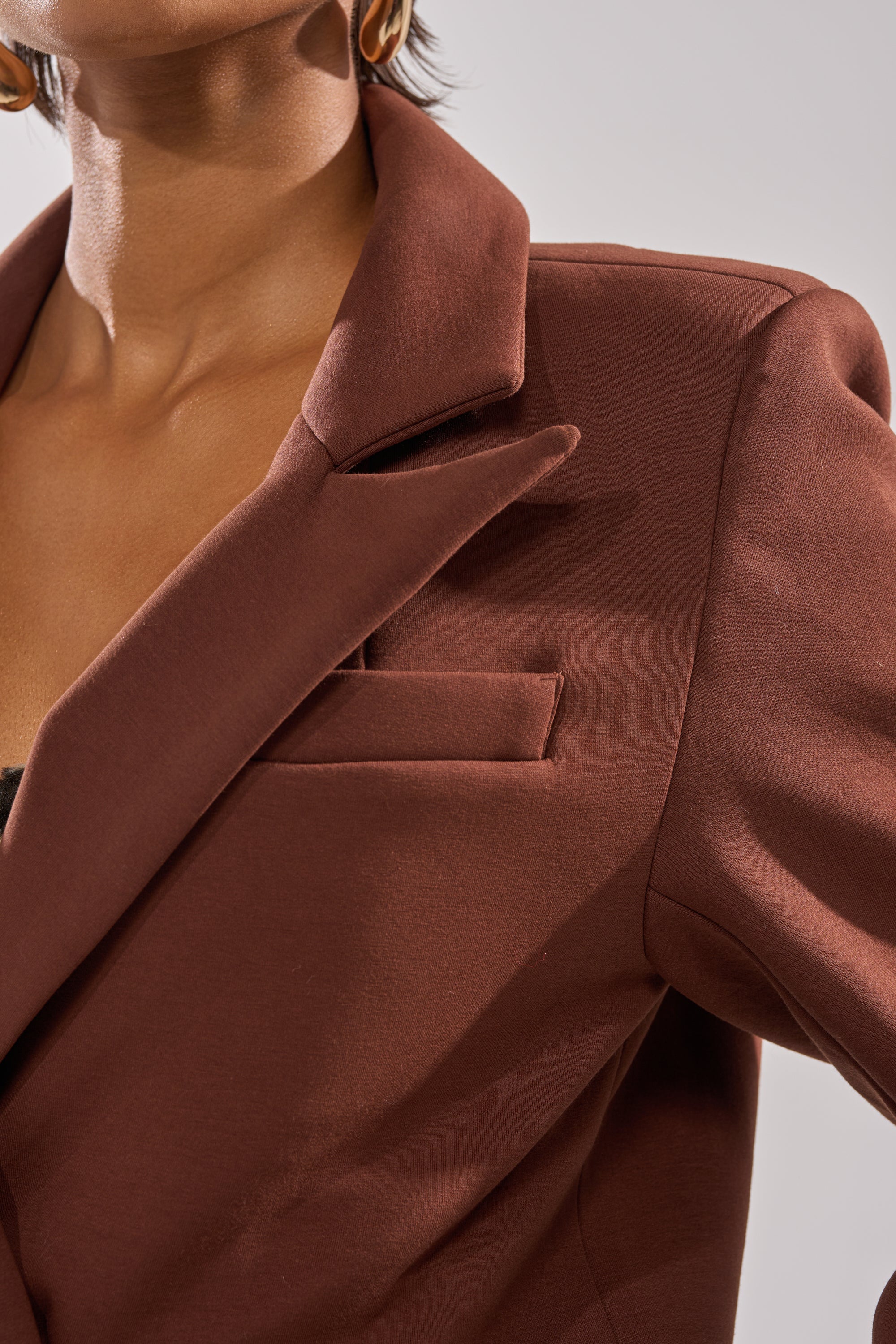 Close-up of a person in the COCOA BUTTER SCUBA TRENCH IN BROWN featuring a notched lapel and chest pocket. A gold hoop earring adds detail against a neutral background, highlighting the sleek style of this brown blazer. 5