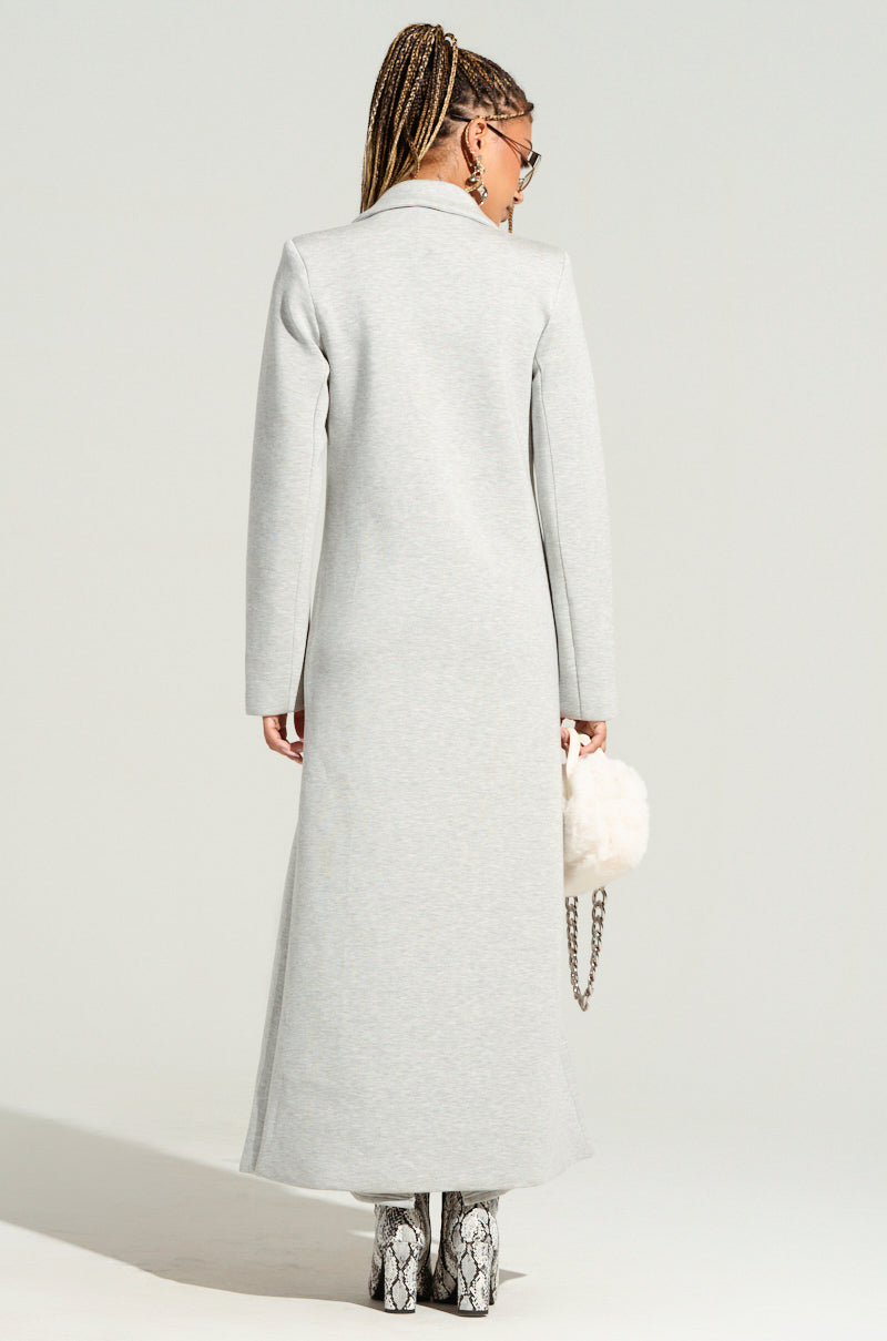 A person with braided hair, wearing the chic COCOA BUTTER SCUBA TRENCH in grey and snakeskin-patterned boots, holds a fluffy white handbag. The coat's double-breasted front enhances their sophistication as they face away from the camera against a plain, light-colored background.