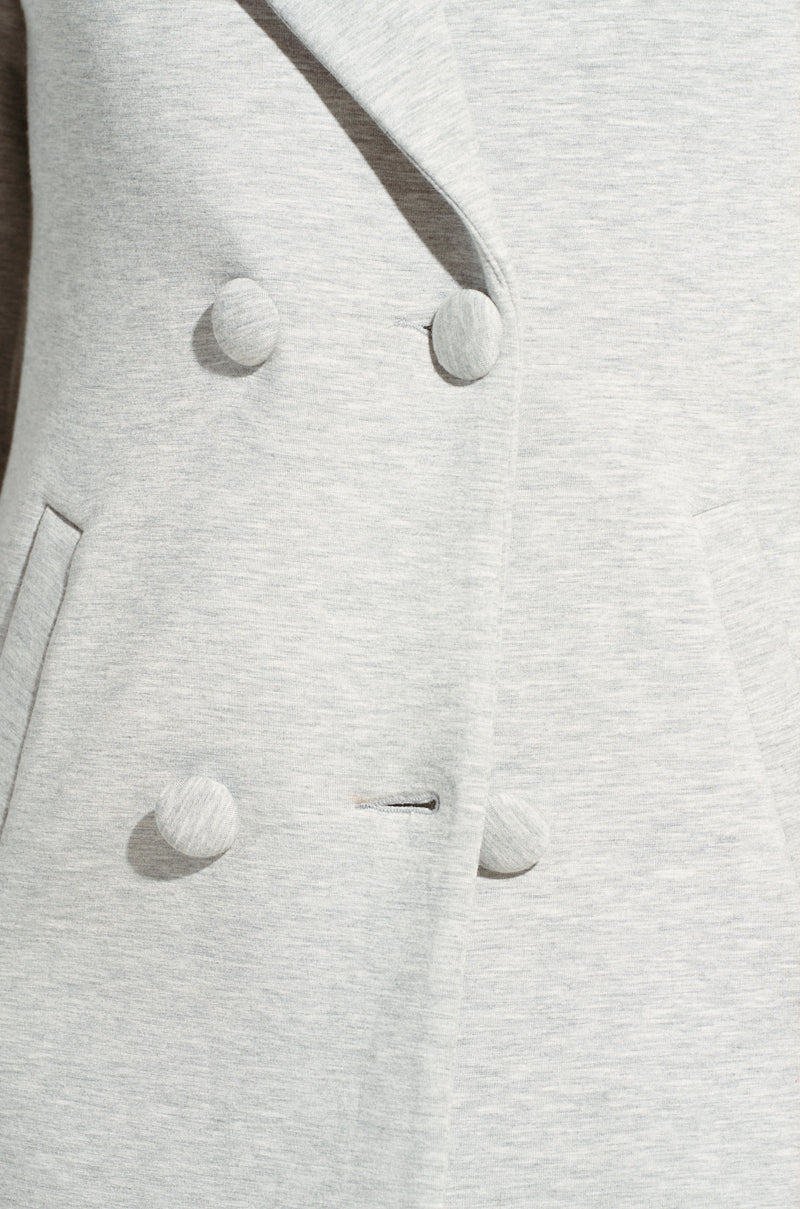 Close-up of the COCOA BUTTER SCUBA TRENCH IN GREY with a double-breasted front. This gray trench showcases round buttons and two visible pockets on either side, emphasizing its texture and tailoring details reminiscent of a classic wool blazer style.