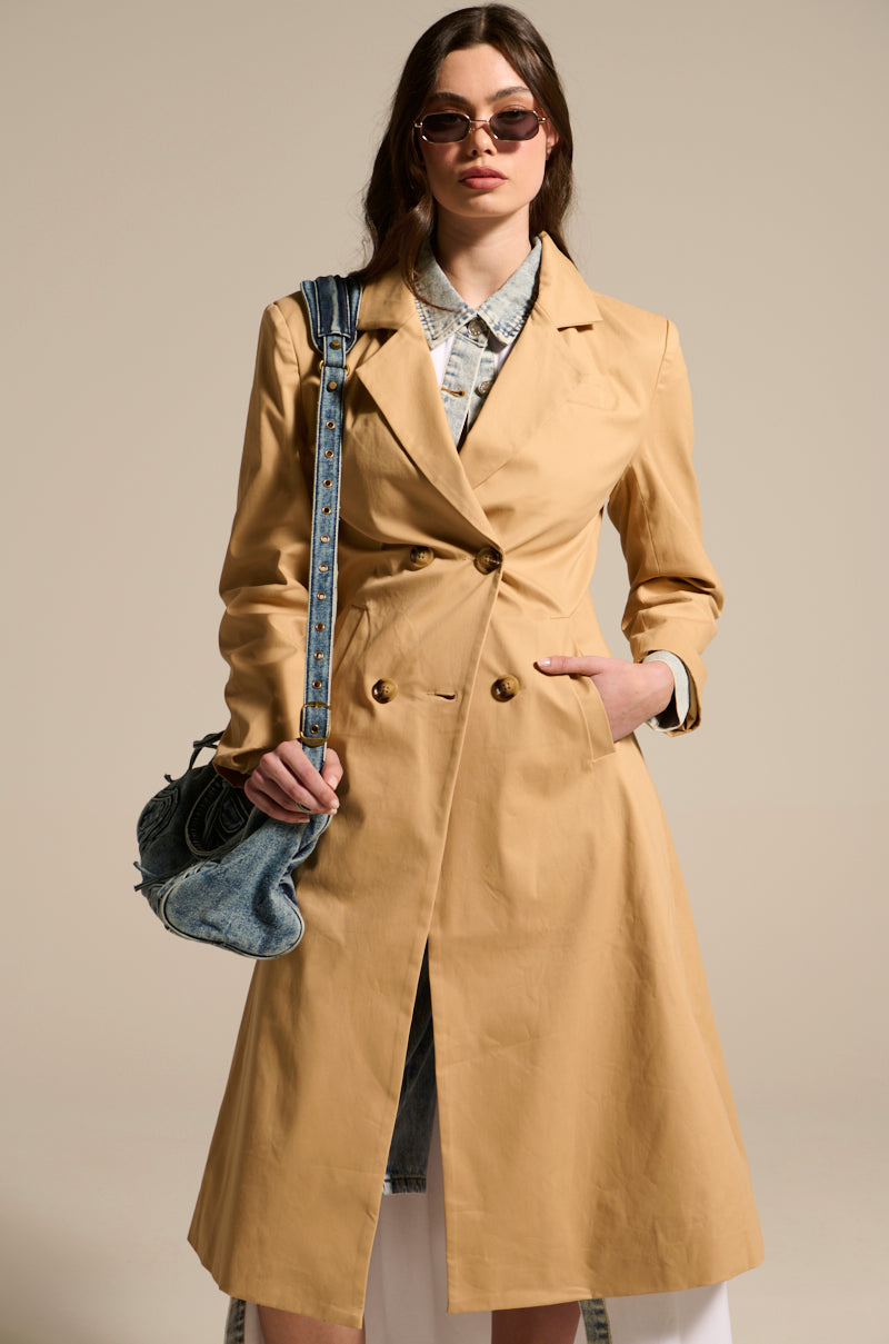 A person confidently wears khaki jade-structured trench coat, sunglasses, and a denim shirt underneath. They carry a blue denim handbag slung over their shoulder against a plain, neutral background.