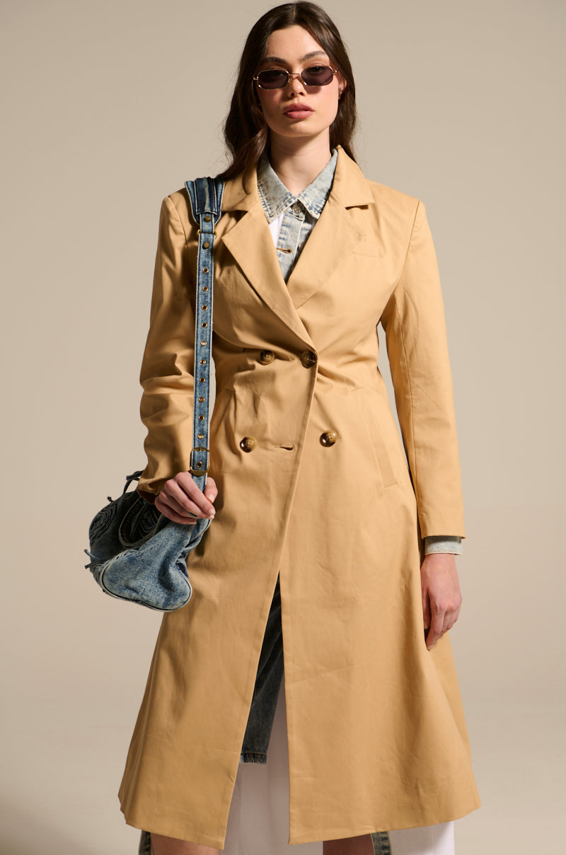 A person wears the JADE STRUCTURED TRENCH IN KHAKI over a denim shirt and skirt, accessorized with sunglasses and a blue denim bag, against a plain background.