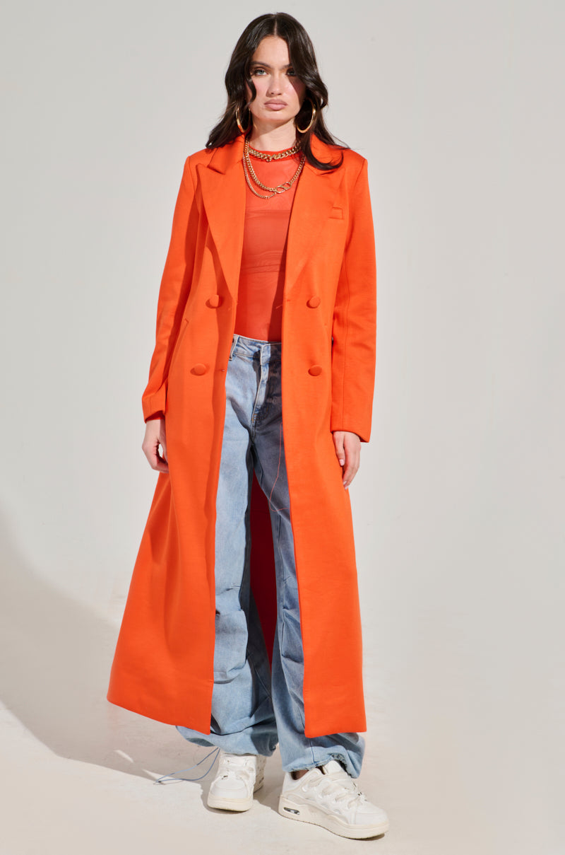 A person stands against a plain background, wearing the bold COCOA BUTTER SCUBA TRENCH IN ORANGE over an orange top, paired with light blue wide-leg jeans and white sneakers. They have long dark hair and are accessorized with a necklace and hoop earrings.