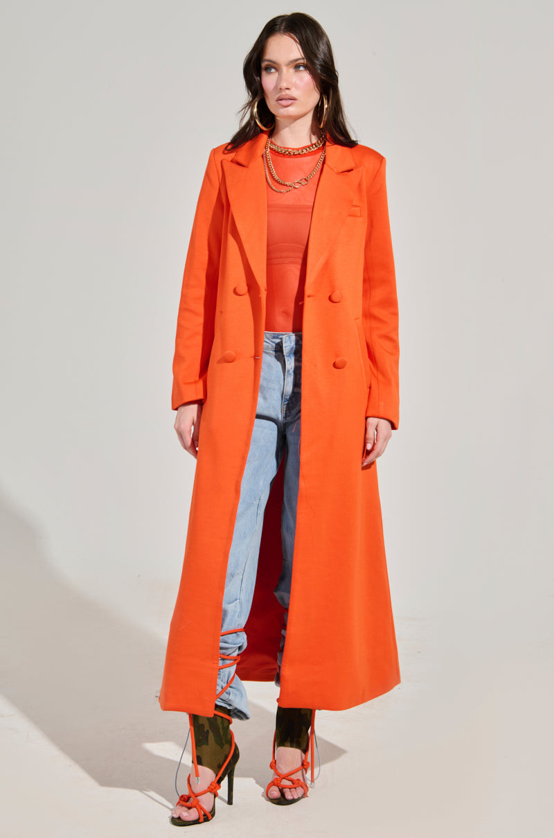 A person wearing the vibrant COCOA BUTTER SCUBA TRENCH IN ORANGE over a matching top, light blue jeans, and orange high-heeled sandals. The outfit is accessorized with layered necklaces against a plain, well-lit backdrop.