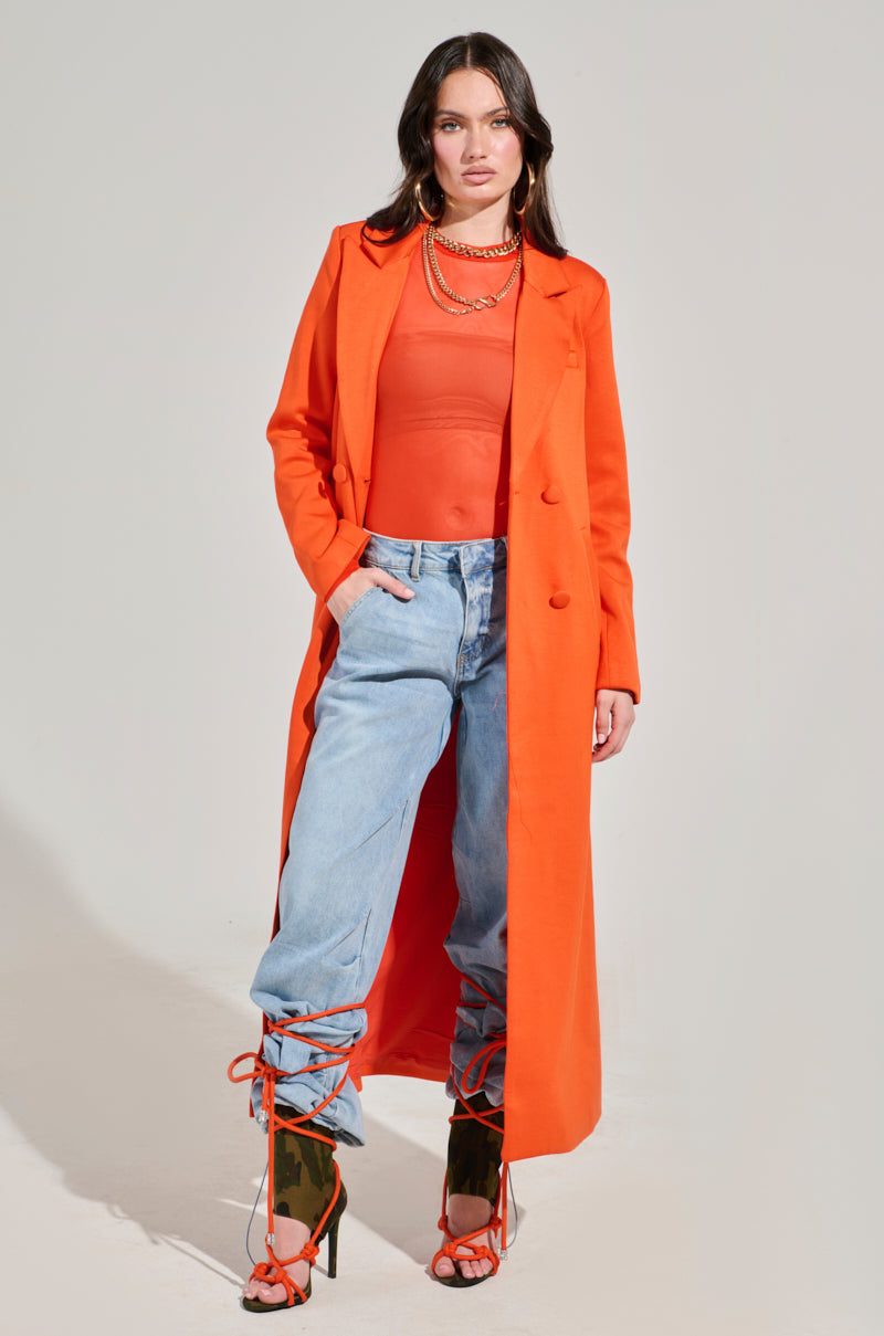 A person stands wearing the COCOA BUTTER SCUBA TRENCH IN ORANGE over a sheer top, paired with light blue jeans. They have strappy orange high heels and layered necklaces, creating a striking look. The background is plain and well-lit.