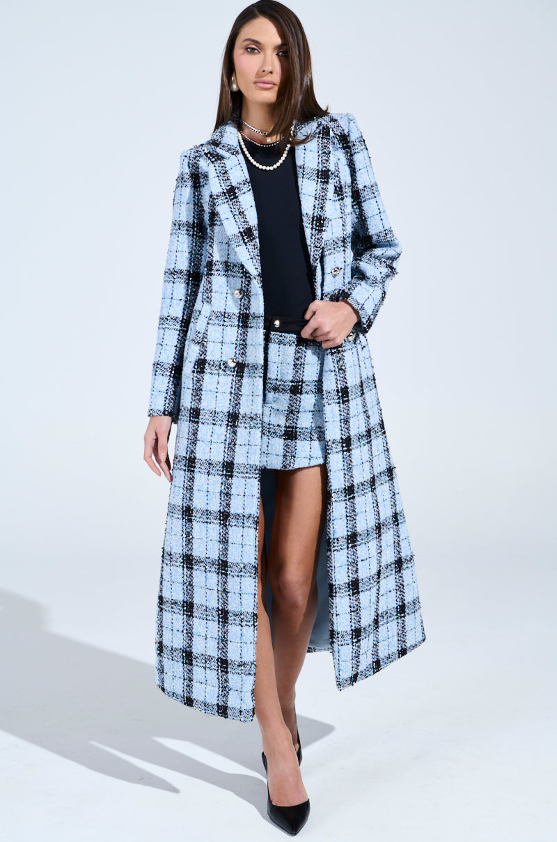 A person stands confidently in a stylish SMOOTH TRANSITION TWEED TRENCH paired with a matching short skirt and black heels. Their long hair complements their pearl necklace against a plain white background.