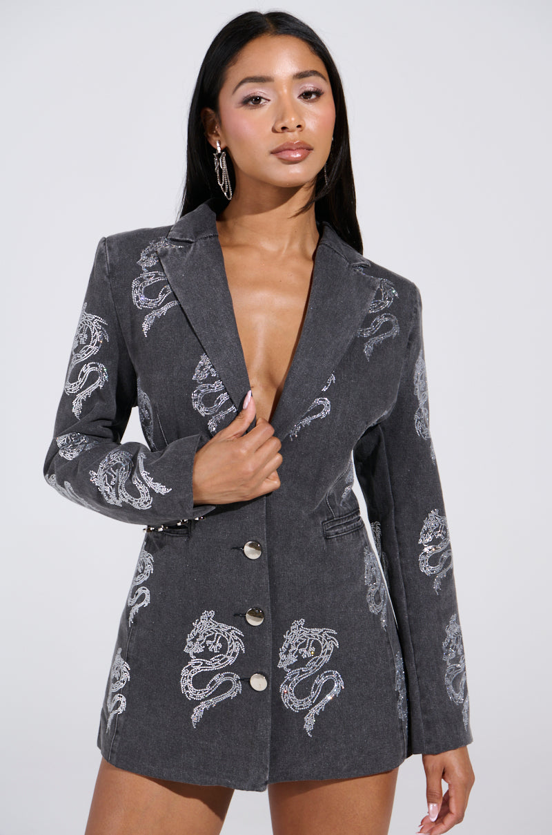 A woman confidently poses in a grey DRAGON SENSE EMBELLISHED BLAZER adorned with intricate dragon patterns. Her long dark hair and large hoop earrings complement the bold, fashion-forward look, wearing the blazer with no visible shirt underneath.