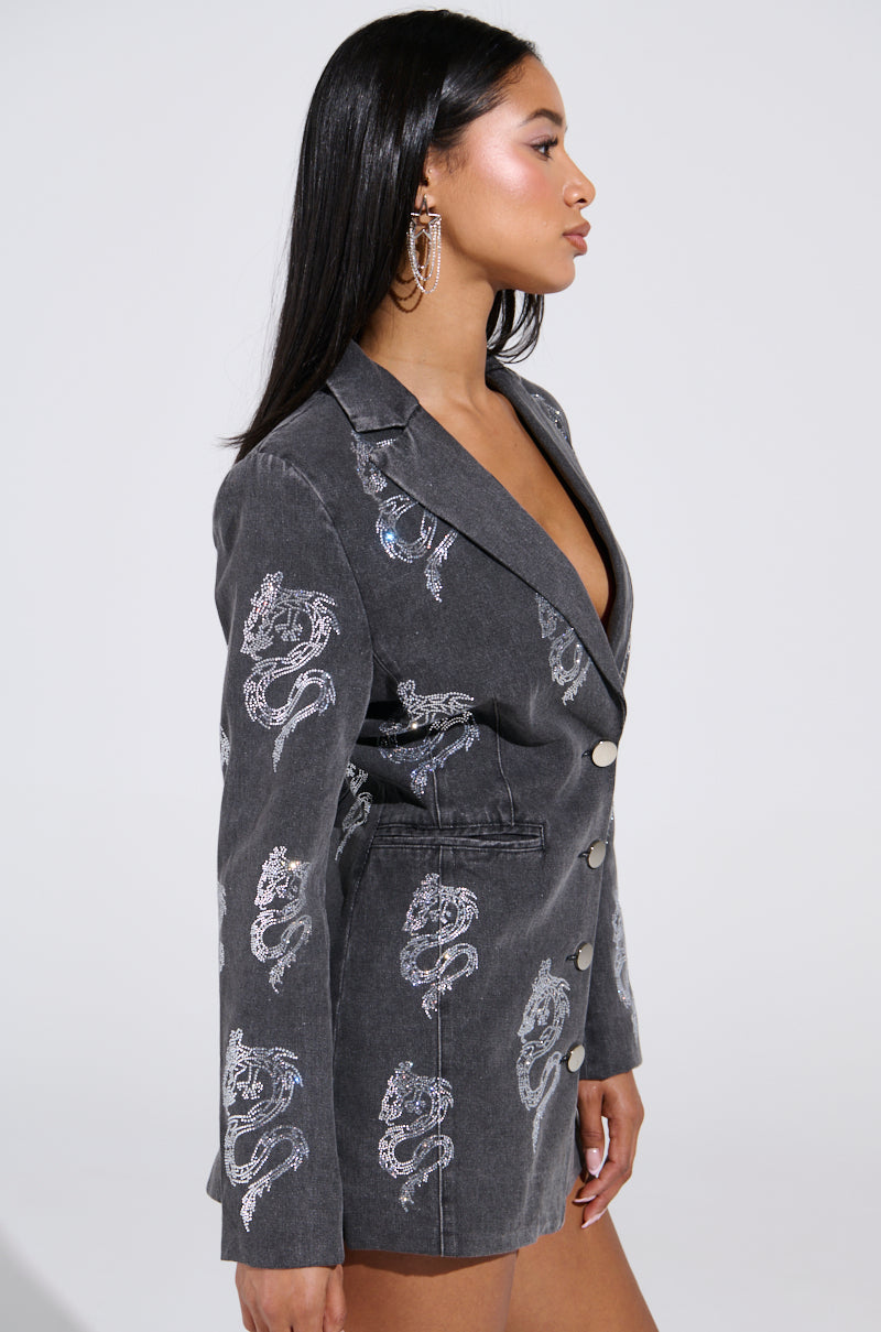 A person with long dark hair is wearing the DRAGON SENSE EMBELLISHED BLAZER, featuring shimmering silver dragon patterns and large buttons. The stylish gray blazer dress sits elegantly over bare shoulders as they stand in profile against a plain white background.