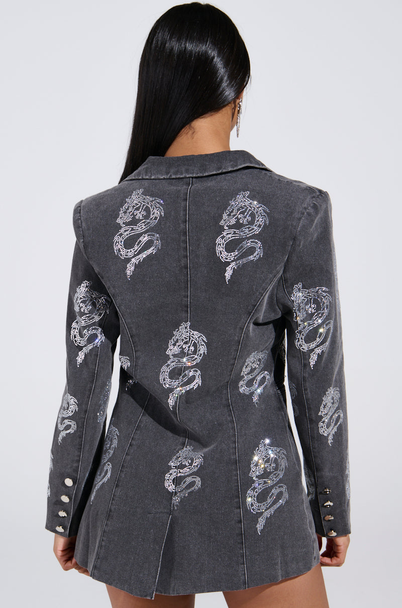 A person with long dark hair wears the DRAGON SENSE EMBELLISHED BLAZER, showcasing shiny dragon designs on the back. The fitted style and buttoned cuffs highlight the intricate rhinestone detailing, adding to its unique charm as they stand facing away.