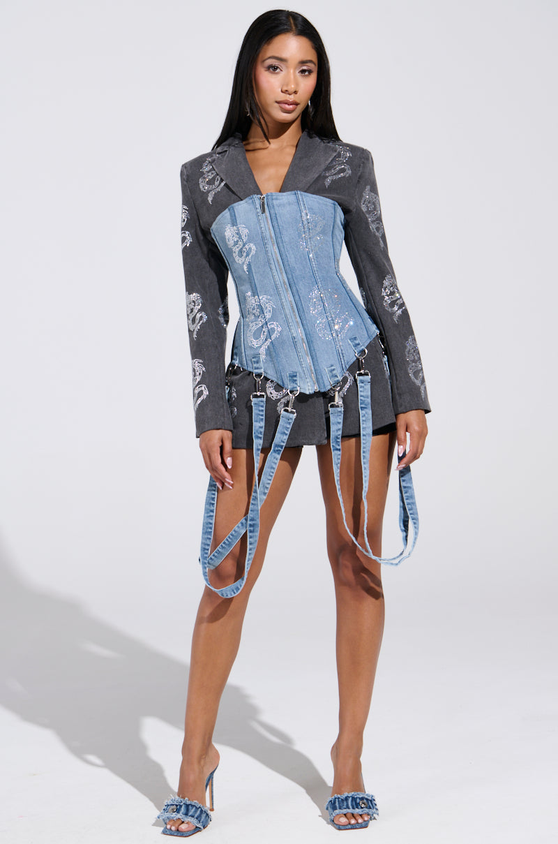 A woman poses against a plain backdrop, wearing a stylish ensemble with a denim corset and garter-like straps. She pairs this look with the Dragon Sense Embellished Blazer, a short skirt, and matching denim heels for an effortlessly chic style.