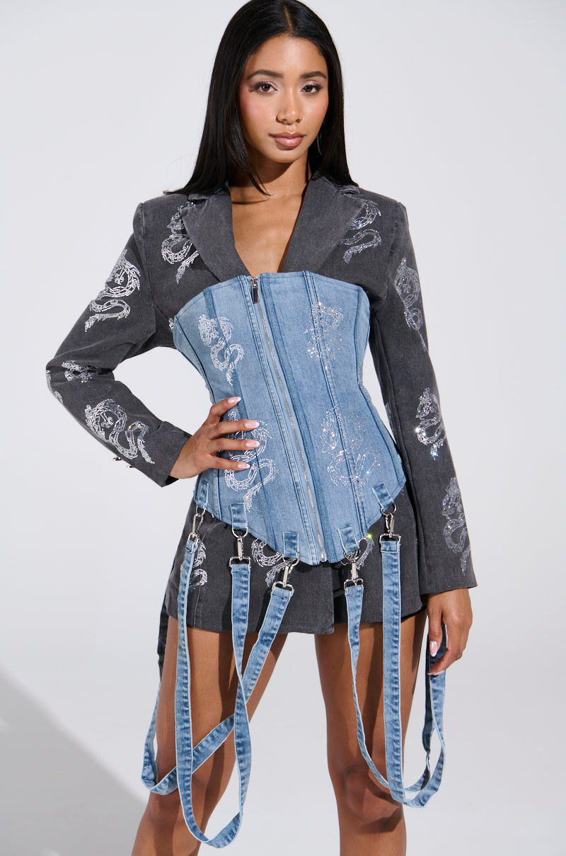 A woman poses confidently in the DRAGON SENSE EMBELLISHED CORSET, a blue denim design with zippers, garter-style straps, and rhinestone details. Her dark jacket is adorned with silver dragon embroidery against a plain backdrop.
