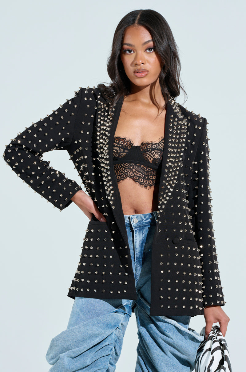With long hair, a person wears the SUGAR AND SPICE SPIKED STUD BLAZER over a lace bralette and high-waisted blue jeans, carrying a black and white patterned bag against a plain backdrop.