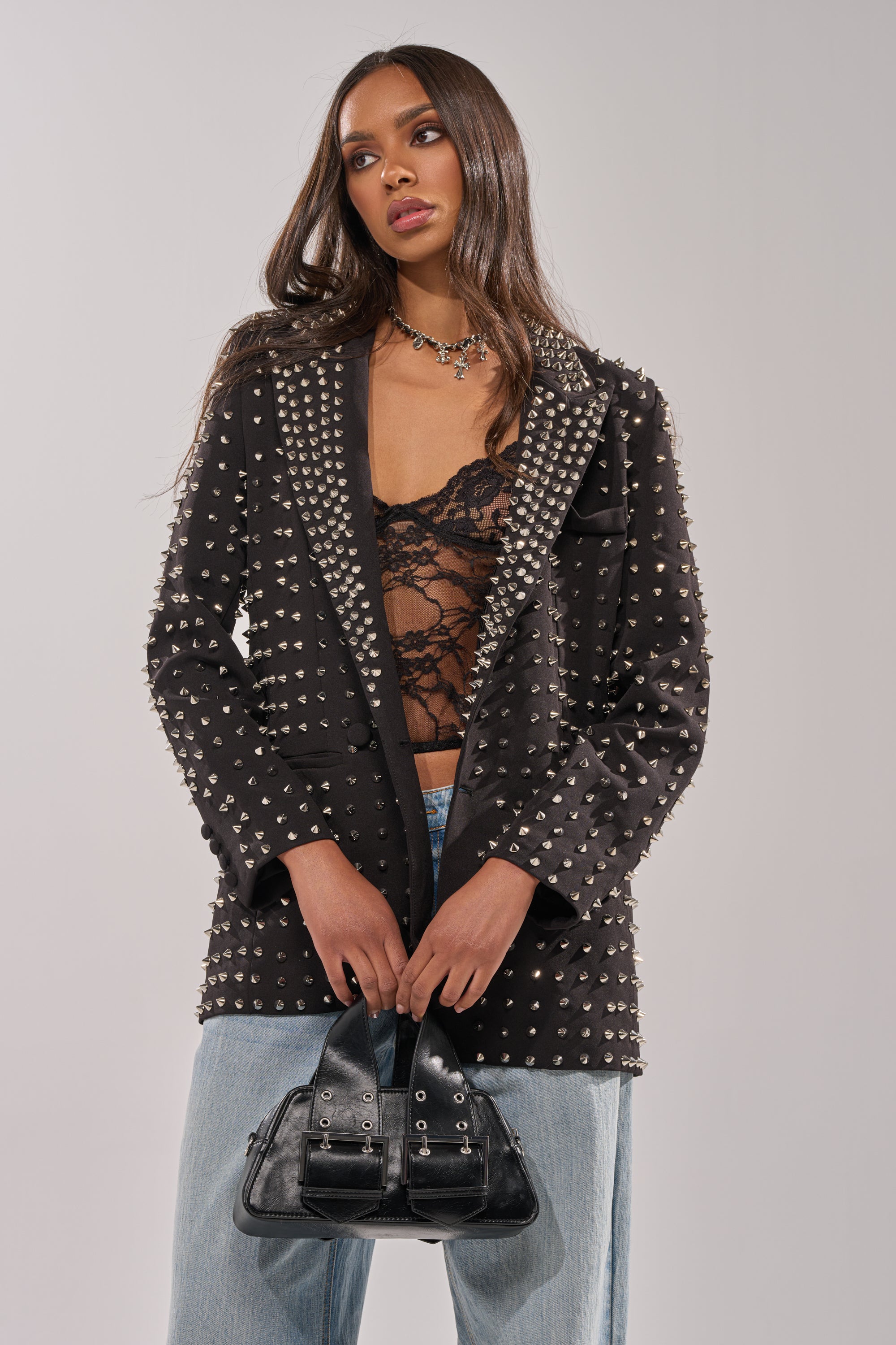 A woman with long brown hair wears the SUGAR AND SPICE SPIKED STUD BLAZER over a black lace top and light blue jeans, holding a small black handbag. She stands against a plain light background, looking off to the side.