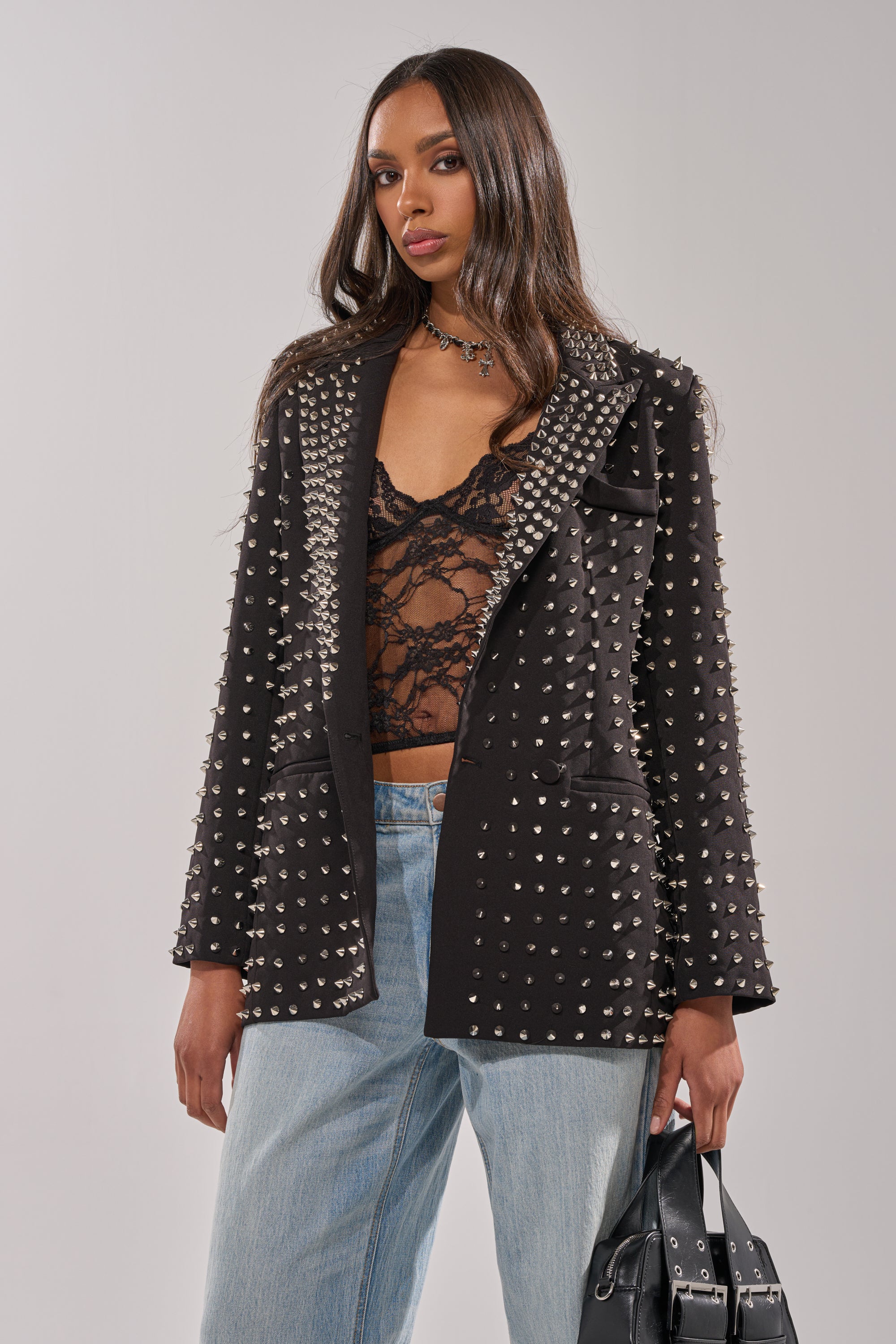 A woman with long brown hair wears the SUGAR AND SPICE SPIKED STUD BLAZER over a sheer black lace top and light blue jeans, holding a black handbag against a plain background.