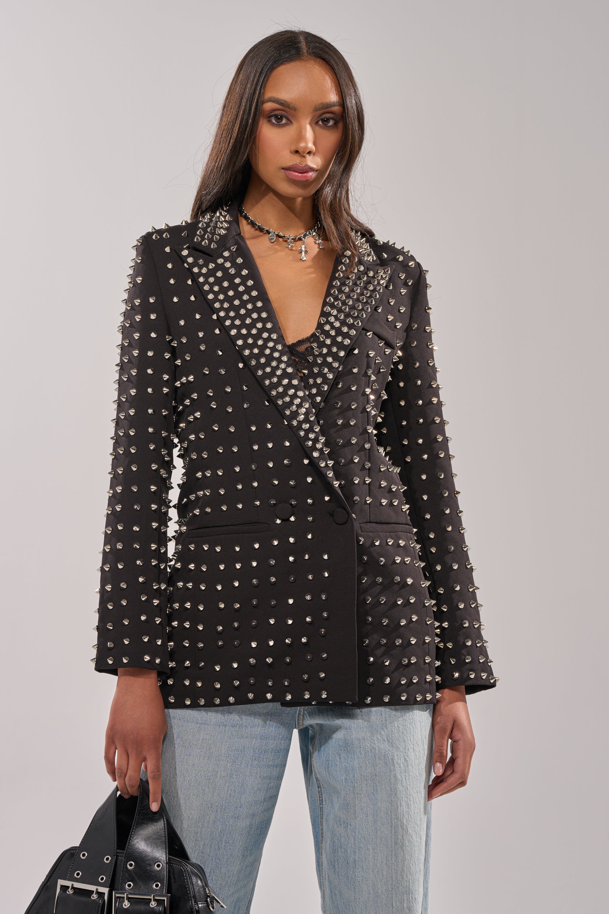 A woman wears the SUGAR AND SPICE SPIKED STUD BLAZER over a lace top and light blue jeans, holding a black handbag. She has long straight hair, a silver necklace, and stands against a plain light background.