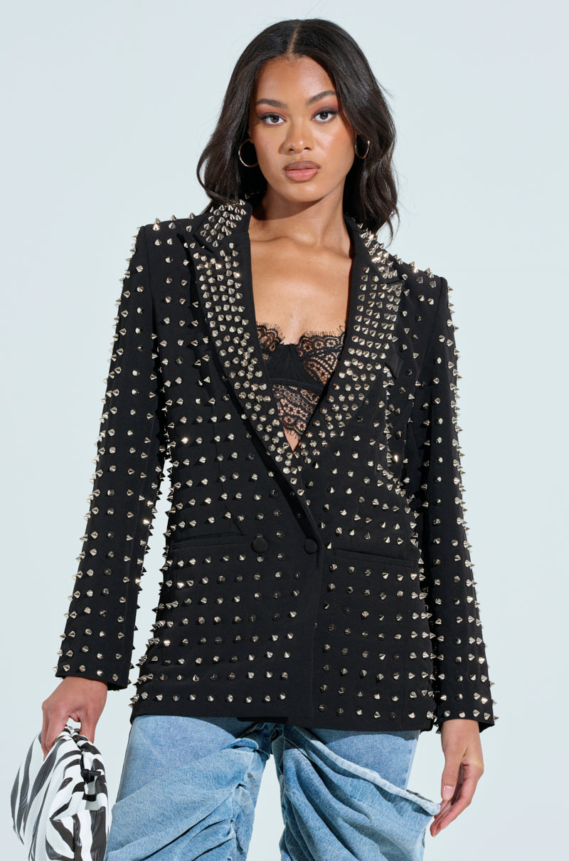 A woman dons the SUGAR AND SPICE SPIKED STUD BLAZER with a notch collar over a lace top, pairing it with light blue jeans. She holds a black and white clutch against a light blue background.
