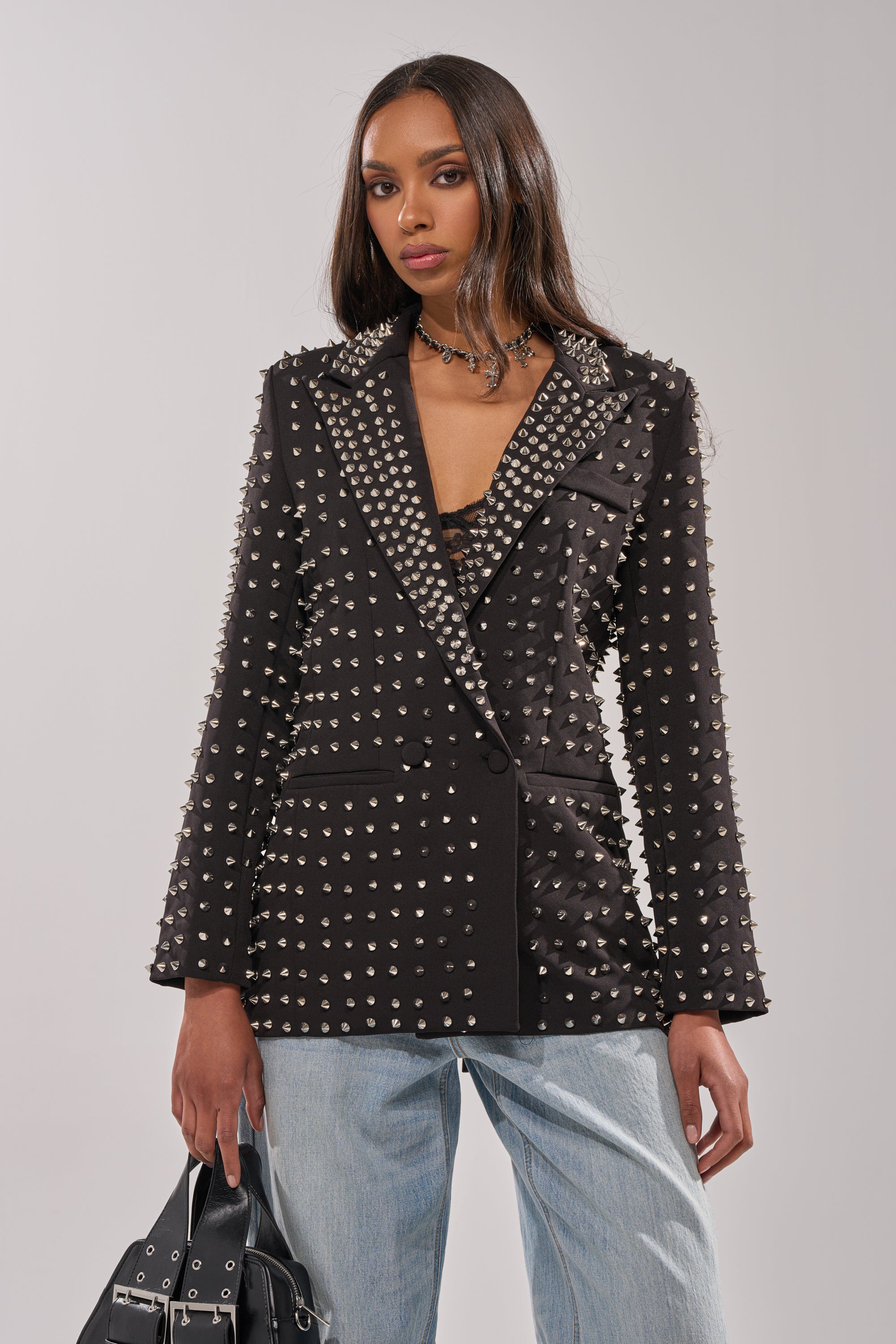 A woman stands against a plain light background wearing the SUGAR AND SPICE SPIKED STUD BLAZER, light blue jeans, and holding a black handbag.
