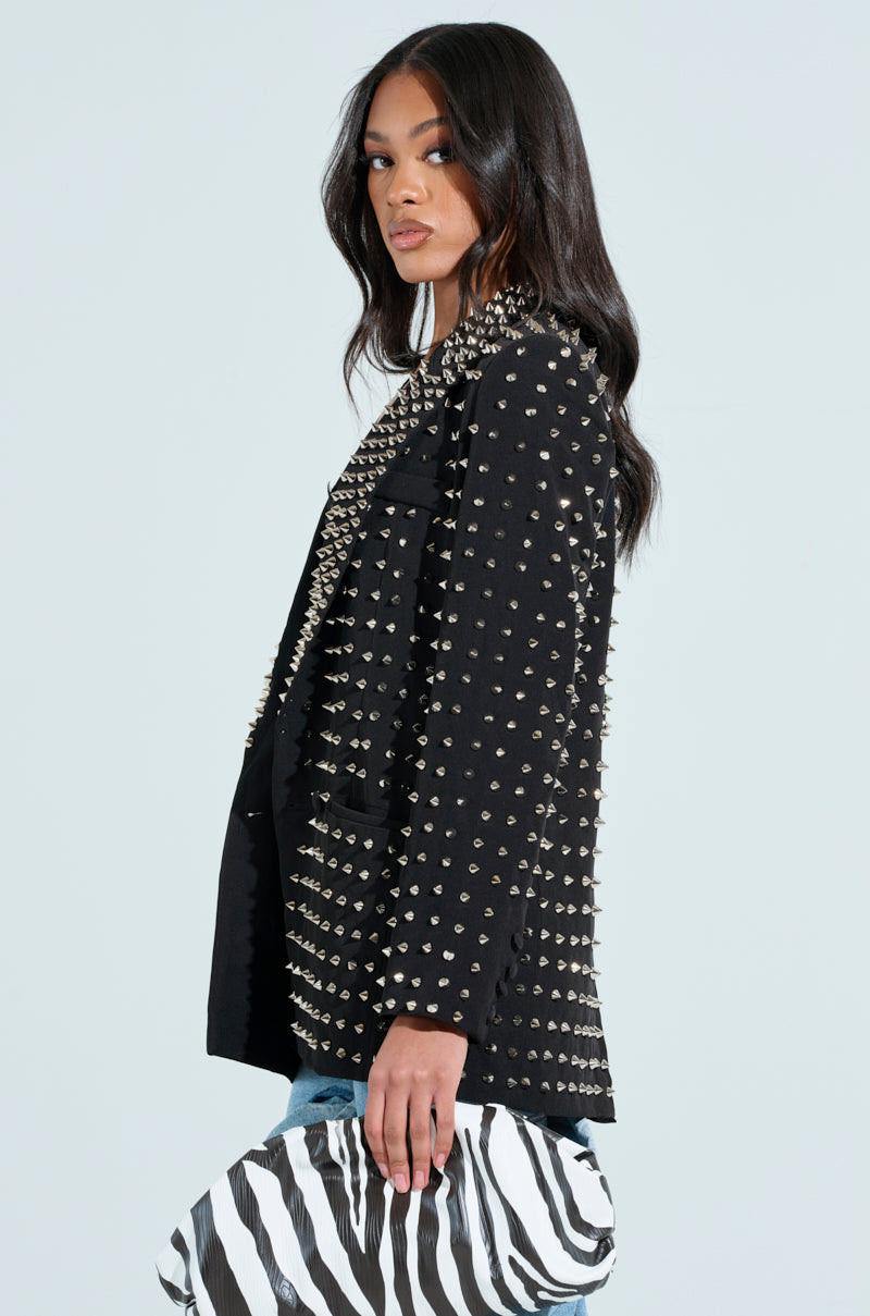A woman in the SUGAR AND SPICE SPIKED STUD BLAZER holds a zebra-striped purse. With long, dark hair, she looks over her shoulder against a plain background.
