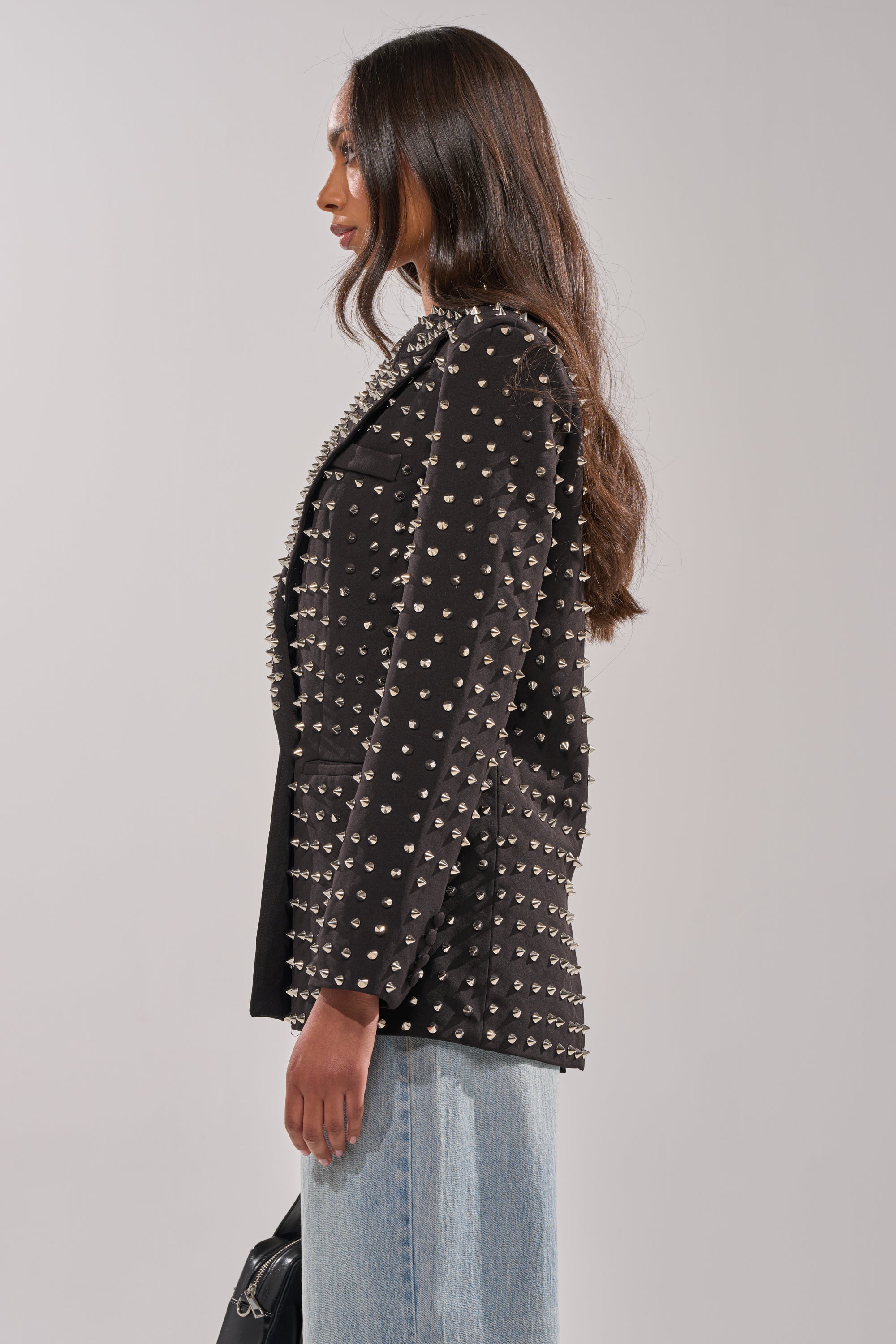 A woman with long brown hair stands in profile wearing the SUGAR AND SPICE SPIKED STUD BLAZER, paired with light blue jeans and a black handbag, set against a plain light background.