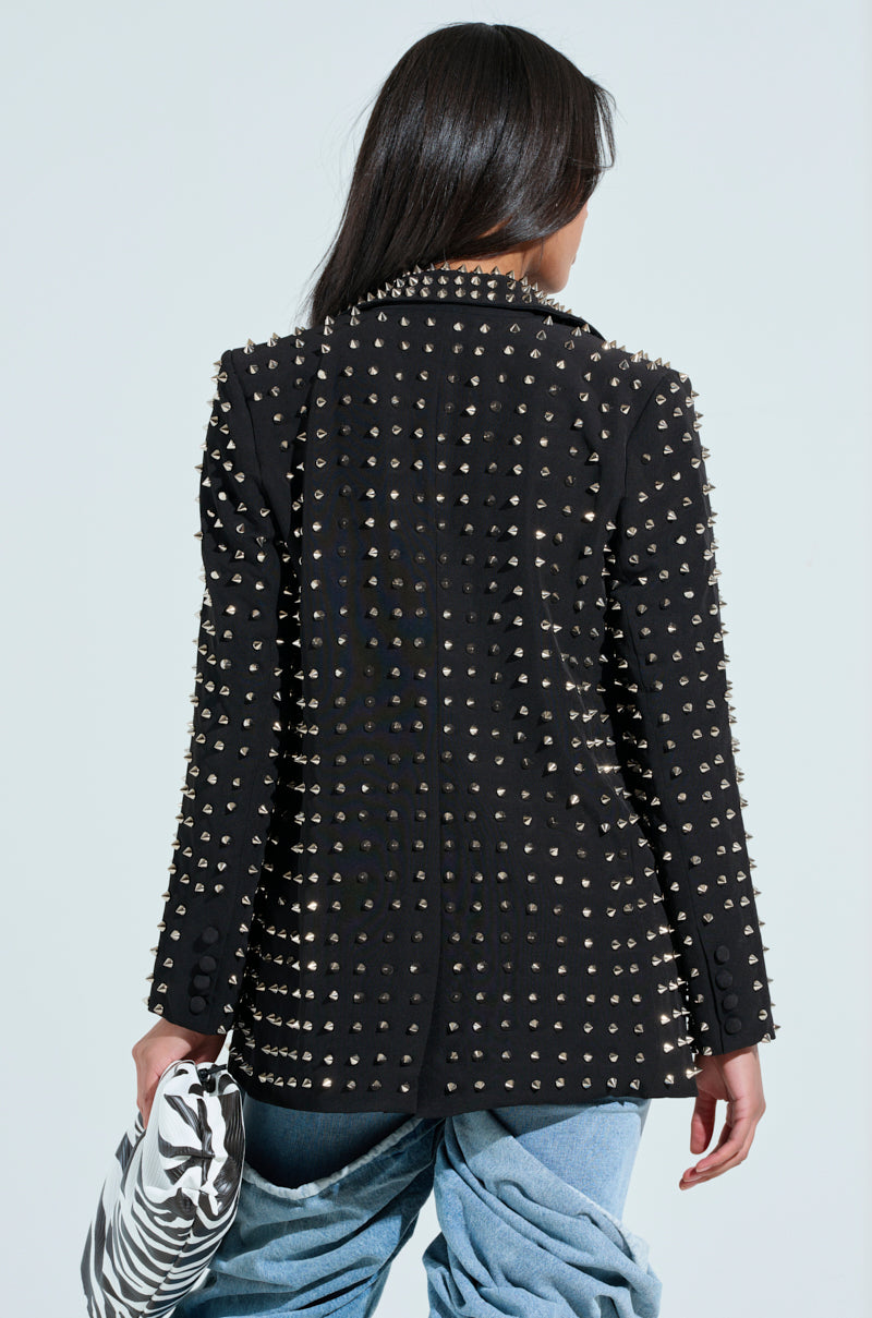 A person with long hair, seen from the back, wears a SUGAR AND SPICE SPIKED STUD BLAZER over blue pants. They hold a zebra-patterned bag in their right hand against a plain background.