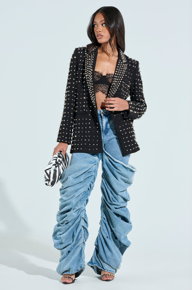 A woman poses in an AKIRA Label SUGAR AND SPICE SPIKED STUD BLAZER over a lace top with baggy ruched denim jeans. She holds a black-and-white patterned clutch, her long wavy hair flowing over black open-toe heels, all against a plain light blue backdrop.