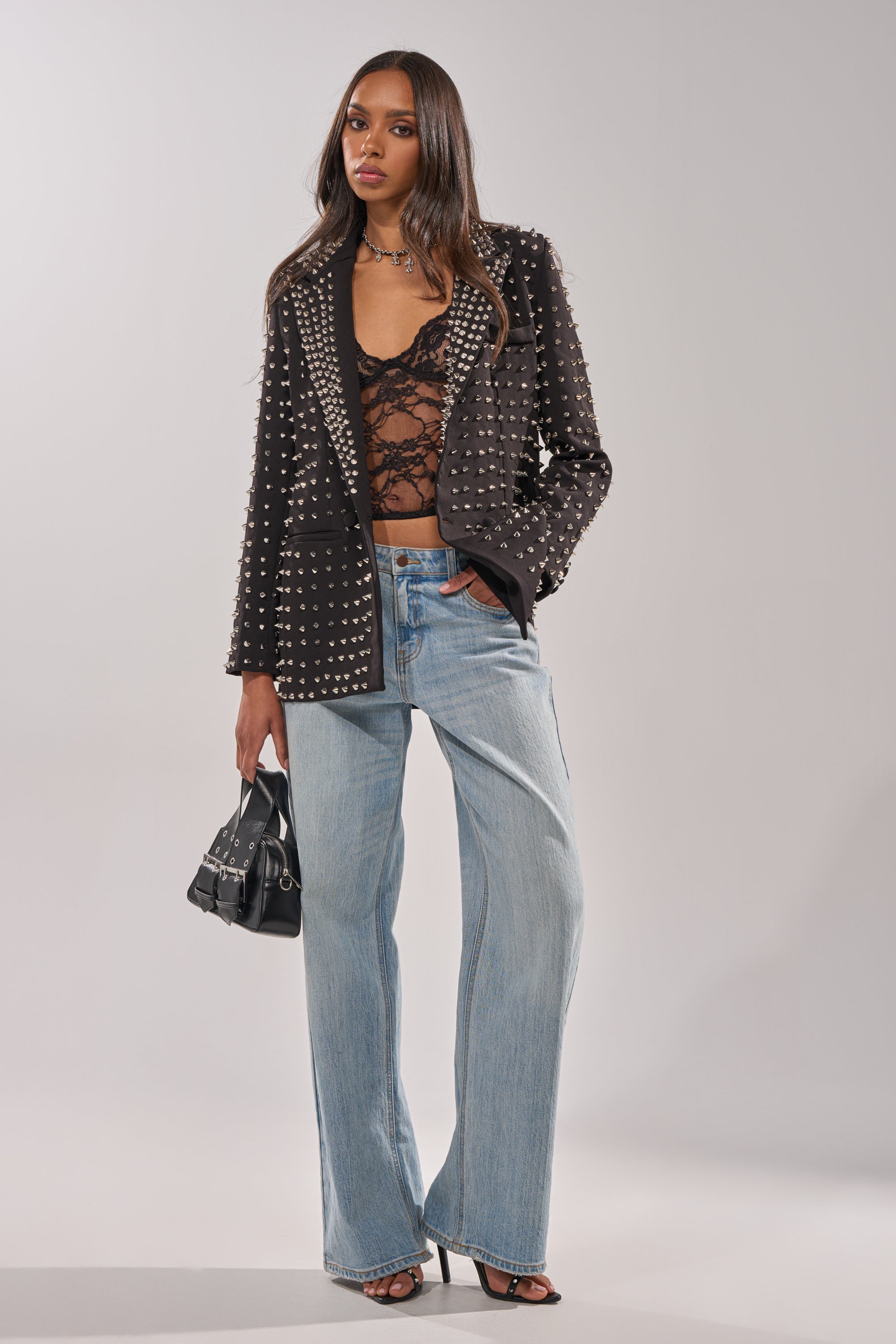 A woman in the SUGAR AND SPICE SPIKED STUD BLAZER over a sheer lace top, light blue wide-leg jeans, black heeled sandals, and holding a small black handbag stands against a plain studio background.