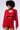 A person with curly hair is wearing a sparkly red outfit from the AKIRA Label, which includes the POP STAR HOT FIX RHINESTONE CROP TOP IN RED, a cropped blazer, and high-waisted shorts. They are looking downwards with a neutral expression against a plain background.