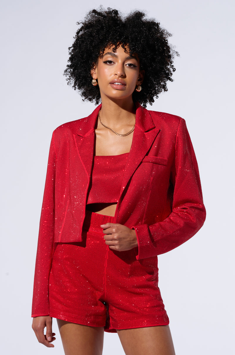 A person with short curly hair is wearing the POP STAR HOT FIX RHINESTONE CROPPED BLAZER IN RED over a matching crop top and high-waisted shorts. They are posing against a white background with a confident expression. 1