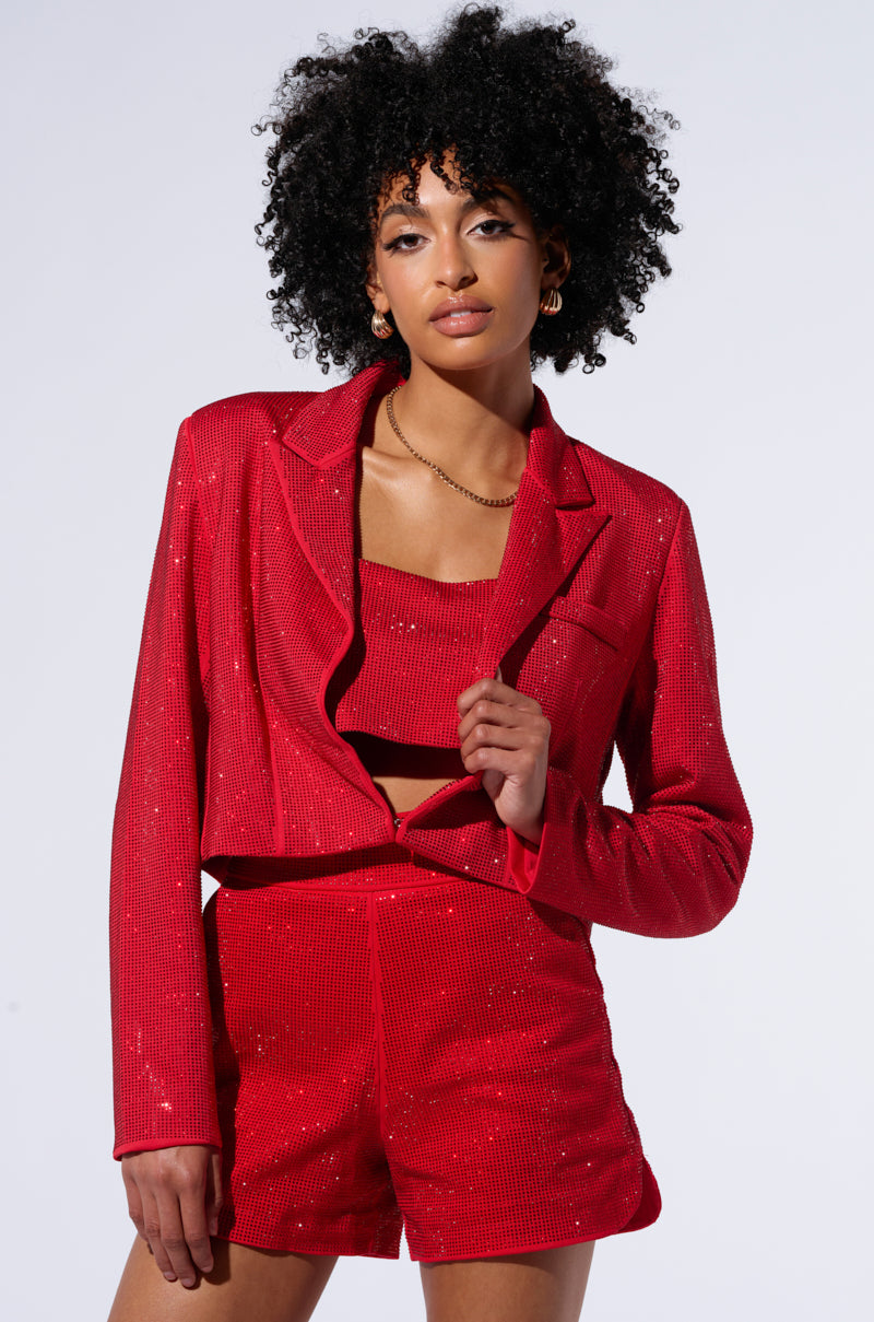 A person with curly black hair stands confidently, wearing the POP STAR HOT FIX RHINESTONE CROPPED BLAZER IN RED from the AKIRA Label, paired with matching high-waisted shorts and a crop top. They hold the lapel of their sparkling red blazer with one hand and have a calm expression on their face. The background is plain and light-colored. 2