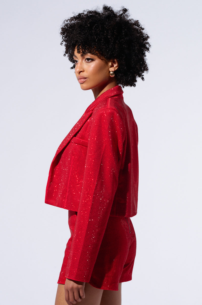 A person with curly hair is confidently posing in the POP STAR HOT FIX RHINESTONE CROPPED BLAZER IN RED and matching shorts from the AKIRA Label. The photo is taken from the side, showcasing the stylish outfit against a plain light background. 4