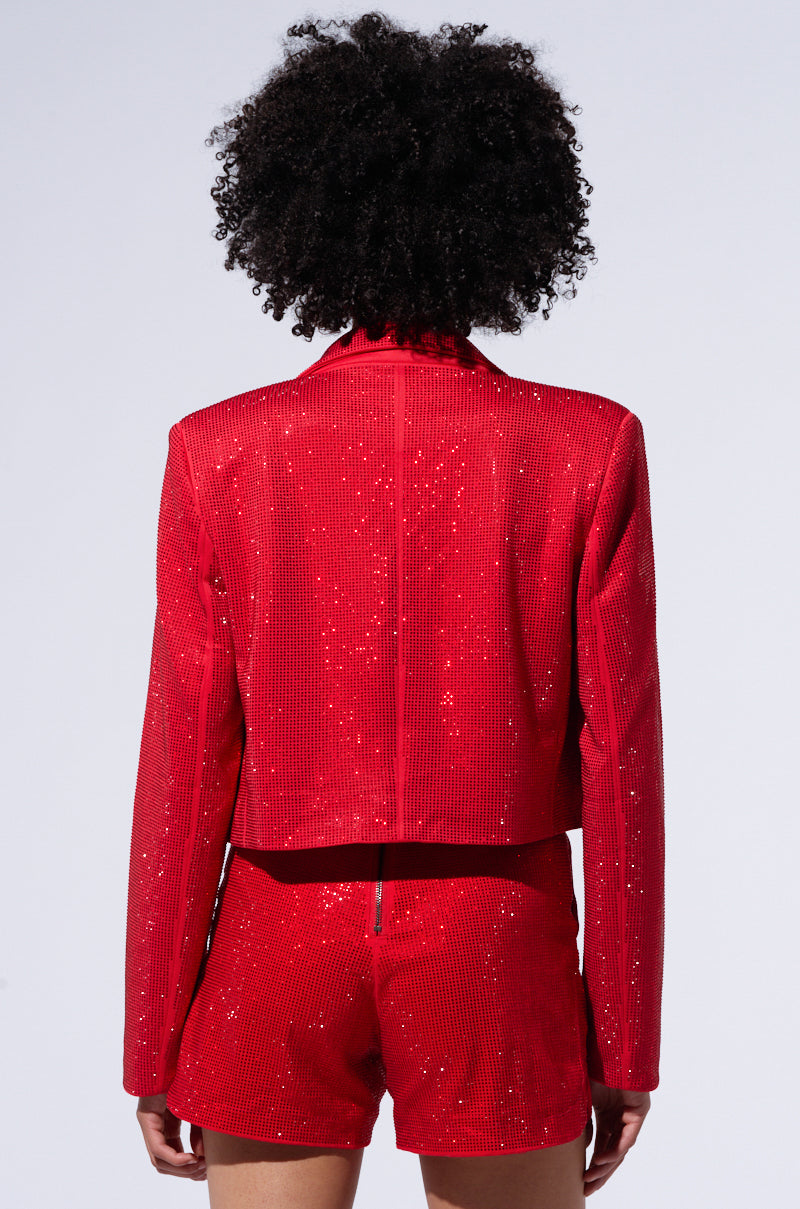 A person with curly hair is facing away from the camera, wearing a sparkly red POP STAR HOT FIX RHINESTONE CROPPED BLAZER IN RED and matching high-waisted shorts. The background is plain and light-colored. 5
