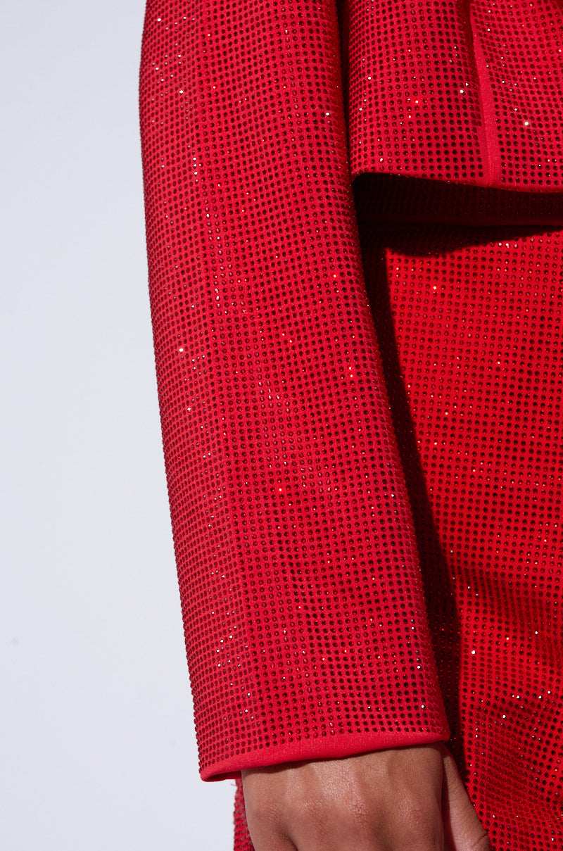 Close-up of a person wearing the POP STAR HOT FIX RHINESTONE CROPPED BLAZER IN RED from the AKIRA Label. The fabric is embellished with a dense pattern of small, glittering rhinestones, giving a shimmering effect. The sleeve is straight with a neat hem, and a portion of the hand is visible at the bottom. 7