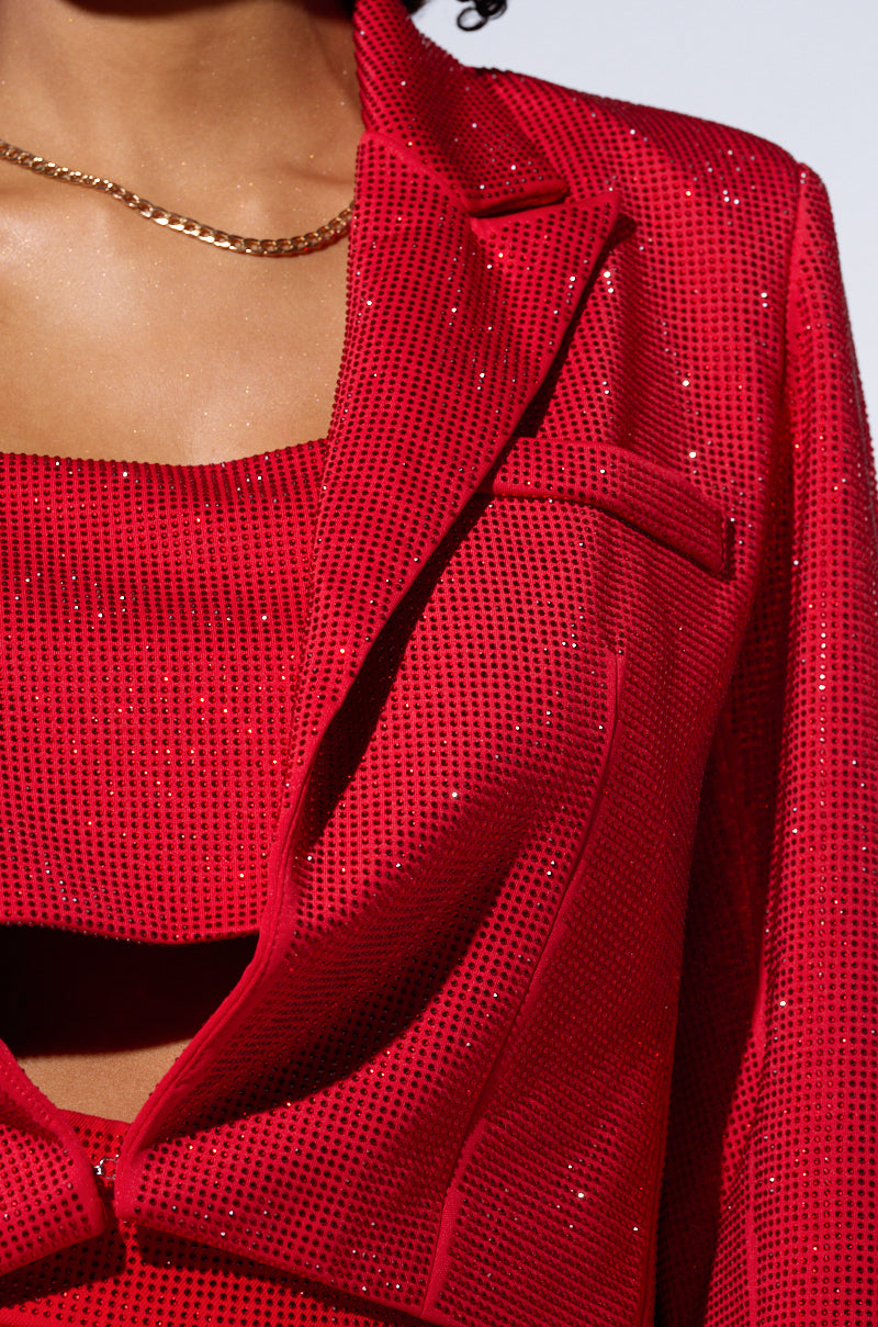 Close-up of a person wearing a sparkling outfit from the AKIRA Label. The ensemble includes the POP STAR HOT FIX RHINESTONE CROPPED BLAZER IN RED and a matching crop top, revealing a chain necklace around their neck. The image captures a partial view, showing part of the face, shoulder, and torso. 8