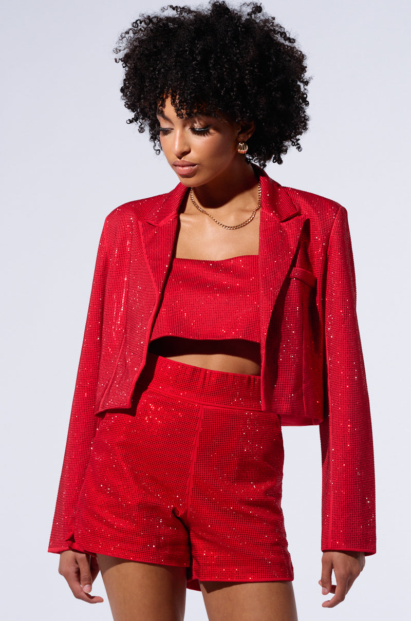 A person with curly hair is wearing a red, glittery outfit consisting of the POP STAR HOT FIX HIGH WAIST RHINESTONE SHORT IN RED paired with a cropped jacket and square-necked crop top. They are looking downward with a serious expression against a plain background.
