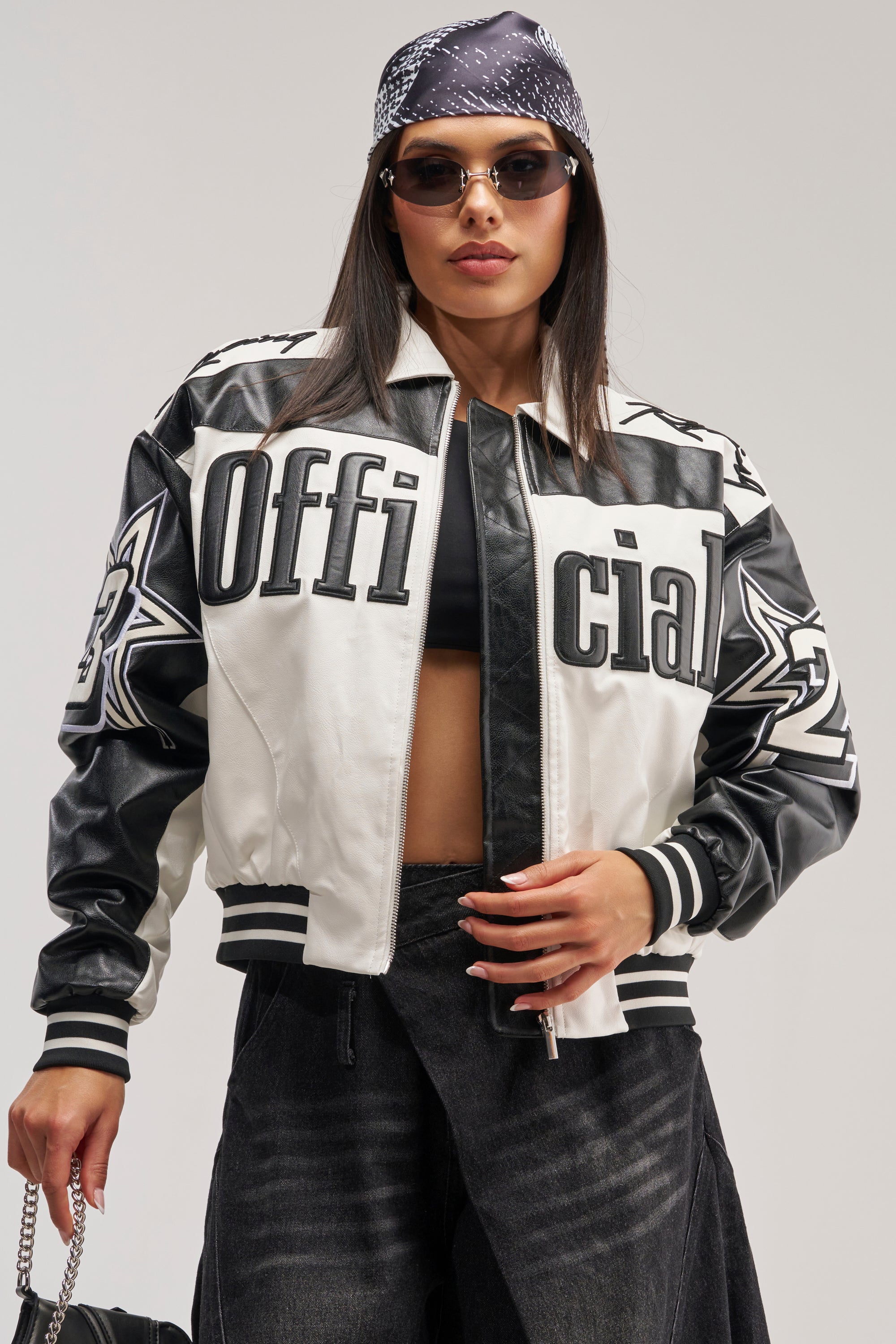 A woman in sunglasses and a black-and-white bandana wears the ON THE RUN MOTO BOMBER, an oversized black-and-white colorblock jacket featuring “Official” on the front. She pairs it with black jeans, carries a chain-strap bag, and poses confidently against a plain background.