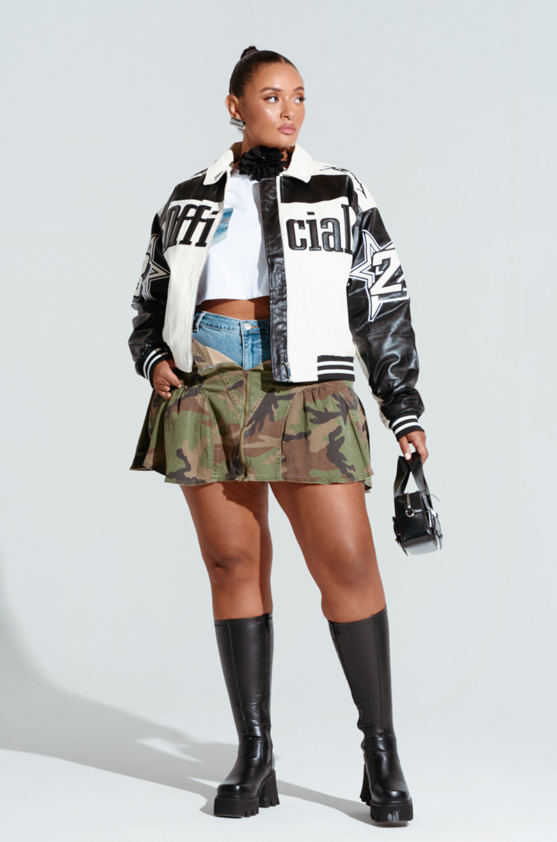 A woman models a stylish outfit featuring the ON THE RUN MOTO BOMBER, a black and white varsity jacket with color block construction. She pairs it with a white top, denim shorts, and a camouflage pleated skirt. Her look is completed with tall black boots and a small black handbag, while her hair is pulled back in a sleek bun.