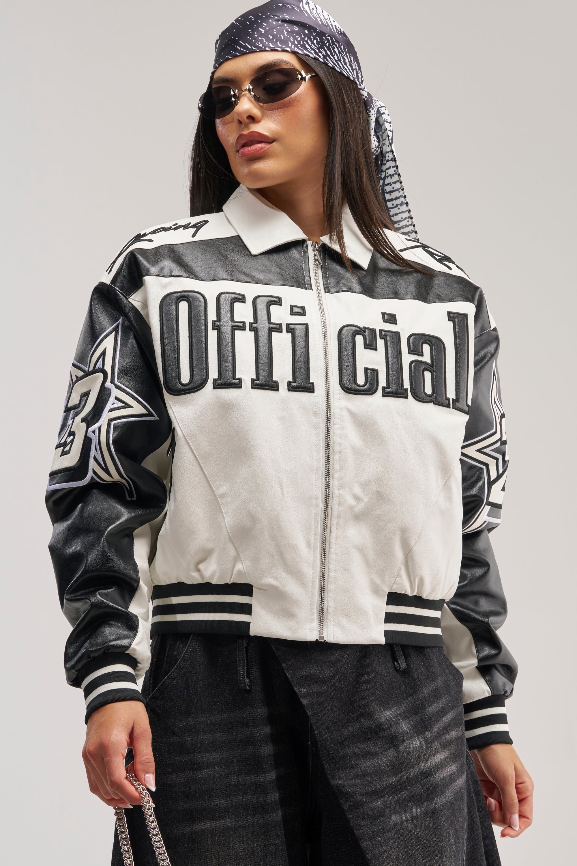 Wearing the ON THE RUN MOTO BOMBER—a racing jacket featuring "Official" on the front and star designs on the sleeves—along with sunglasses and a patterned headscarf, a person stands confidently against a plain background.
