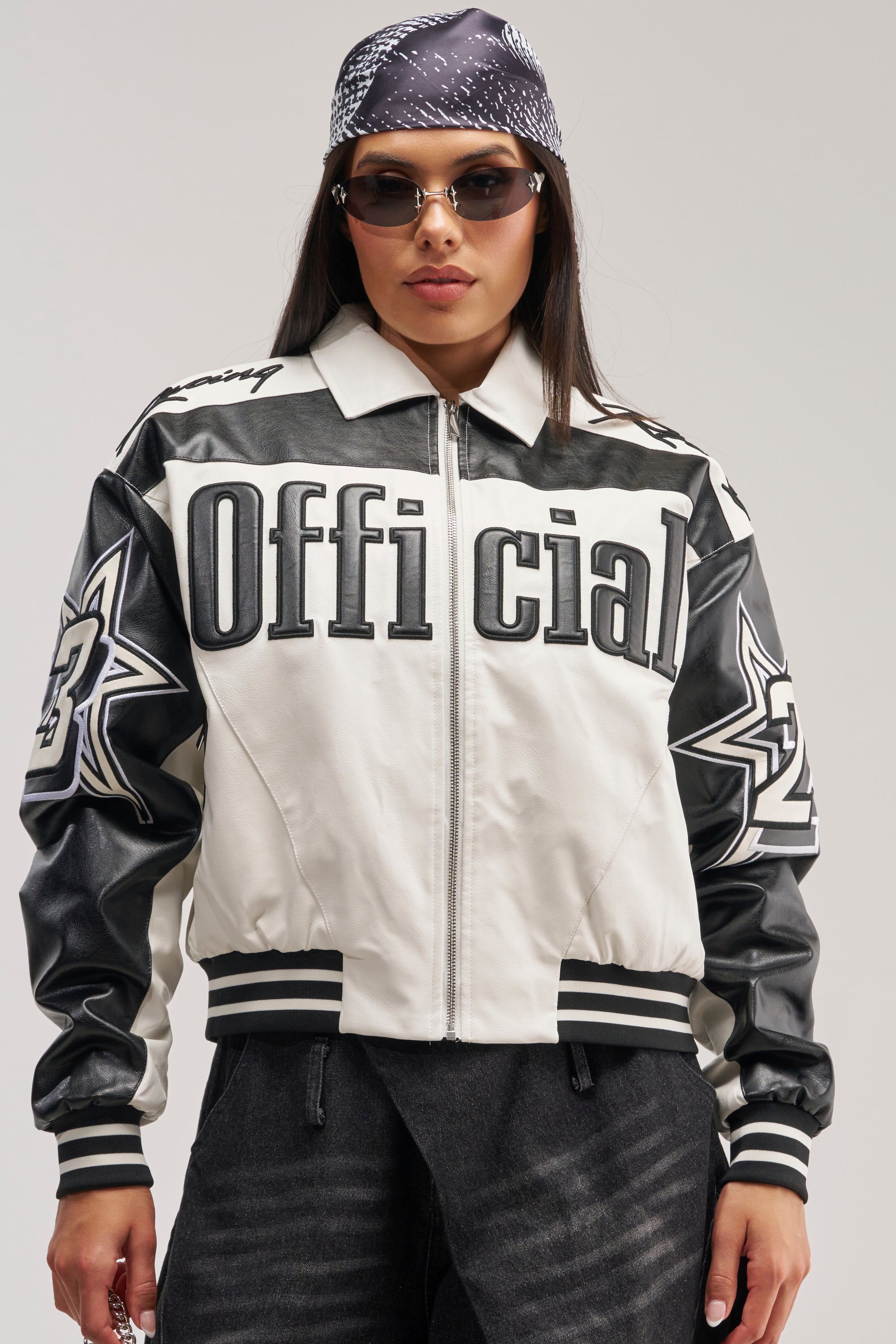 Wearing sunglasses and a bandana, a person stands against a plain background dressed in the ON THE RUN MOTO BOMBER—a black-and-white faux leather jacket featuring "Official" and star designs.
