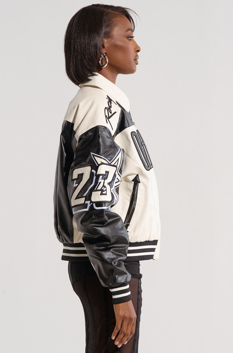 A woman with a short bob hairstyle is wearing the ON THE RUN MOTO BOMBER, a black and white letterman jacket displaying the number "23" and a stylized fox emblem on the sleeve. The racing-inspired embroidered details enhance its sleek design as she stands sideways, facing left.
