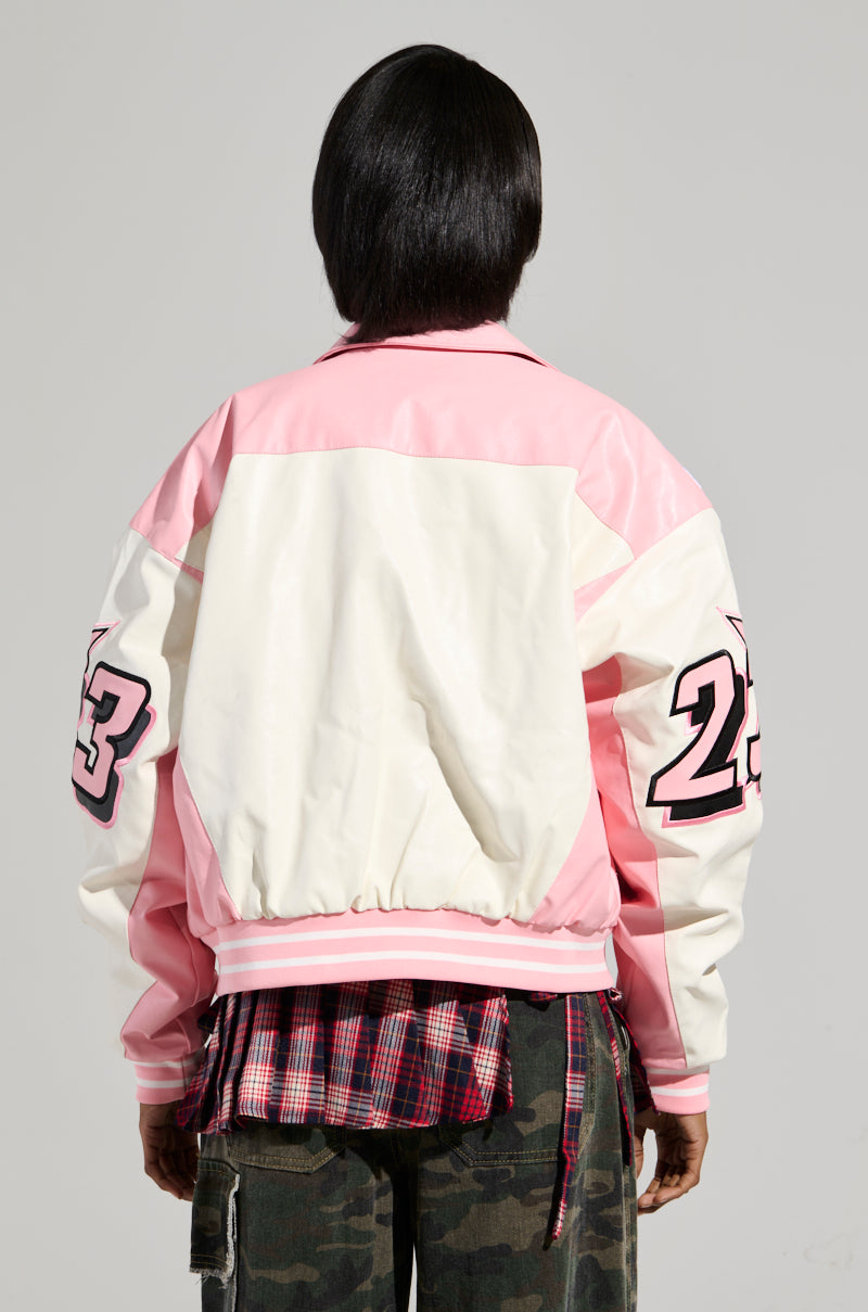 A person stands with their back to the camera, showcasing the KEEP IT MOVING MOTO BOMBER IN LIGHT PINK, which features racing-inspired embroidered patches and the number 23 on the sleeves. The look is completed by a plaid shirt and camouflage pants, creating a unique style fusion.