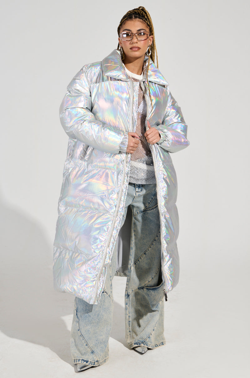 A person stands confidently in the XENON OVERSIZED PUFFER, an iridescent silver coat paired stylishly over a mesh top and distressed wide-leg jeans. They have long braided hair, accessorized with glasses, hoop earrings, and a chain necklace against a plain, bright background.