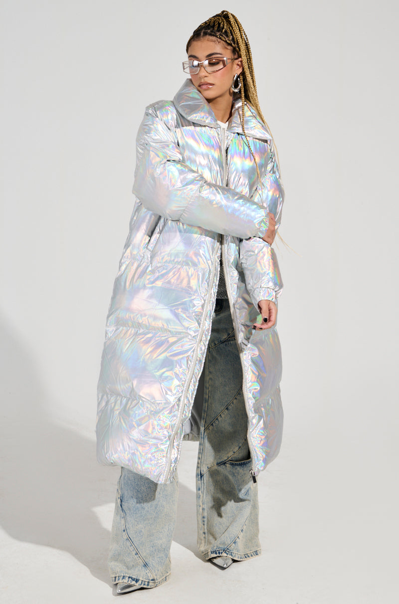 Dressed in the XENON OVERSIZED PUFFER, a person exudes confidence and style against a white backdrop. The iridescent silver coat pairs perfectly with flared jeans, long braided hair, sunglasses, and striking large hoop earrings.