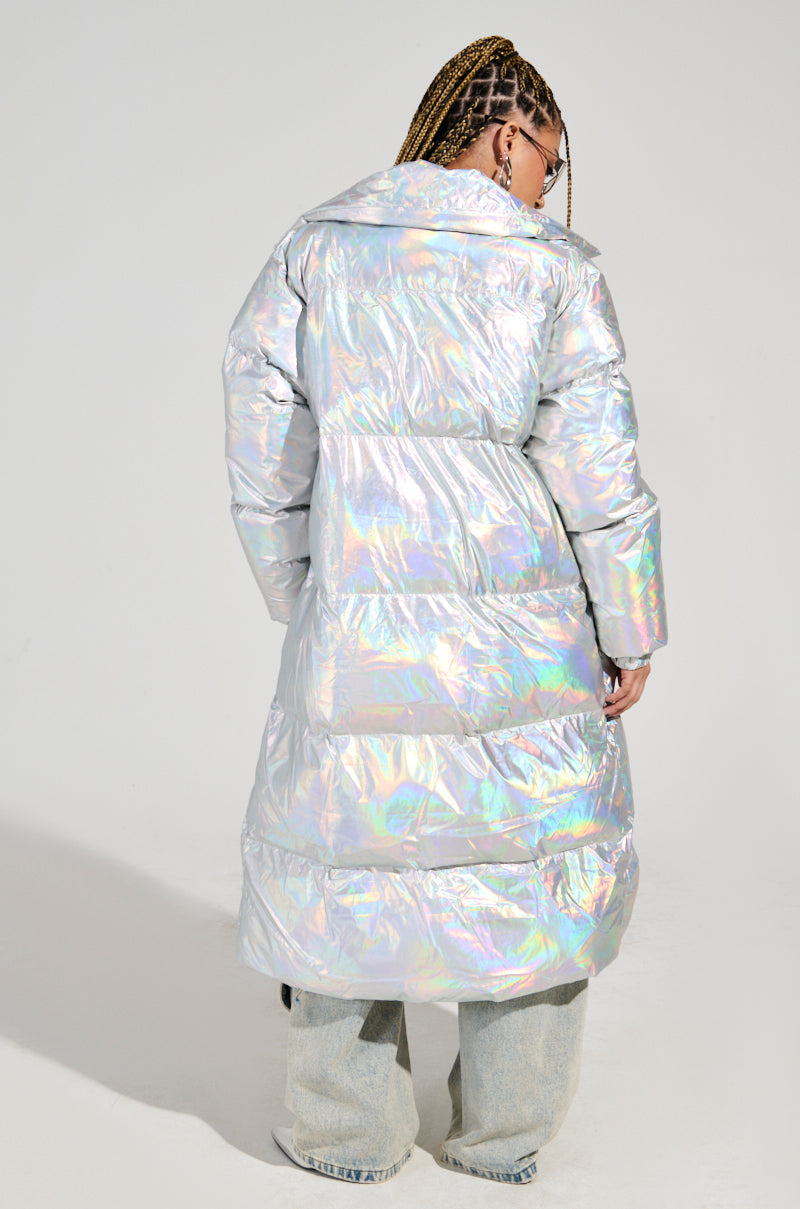 A person with braided hair is wearing the XENON OVERSIZED PUFFER, an iridescent silver coat made of water-resistant polyester. They stylishly pair it with wide-leg jeans and are standing against a plain background, facing away from the camera.
