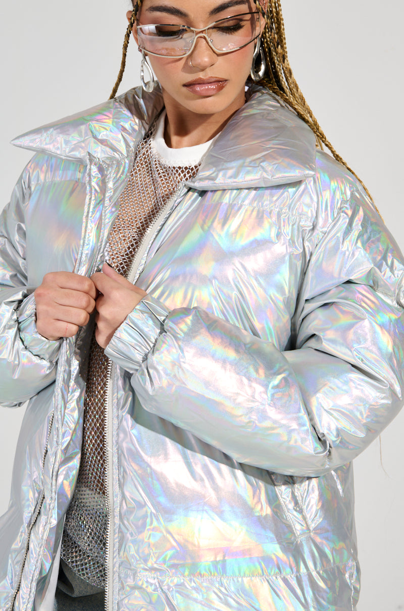 A person wearing the XENON OVERSIZED PUFFER jacket in iridescent silver displays a spectrum of colors under the light. Translucent sunglasses grace their face, and they effortlessly adjust the zipper with confidence. Their braided hair enhances the moment against a plain, neutral background.