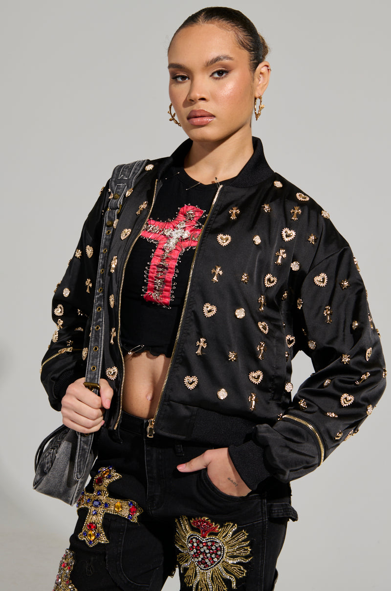 A person poses confidently in the sleek black LIKE A CHARM EMBELLISHED BOMBER, adorned with metallic gold accents, perfectly complemented by a matching outfit featuring cross designs. They carry a gray handbag over one shoulder, set against a plain, light-colored background.