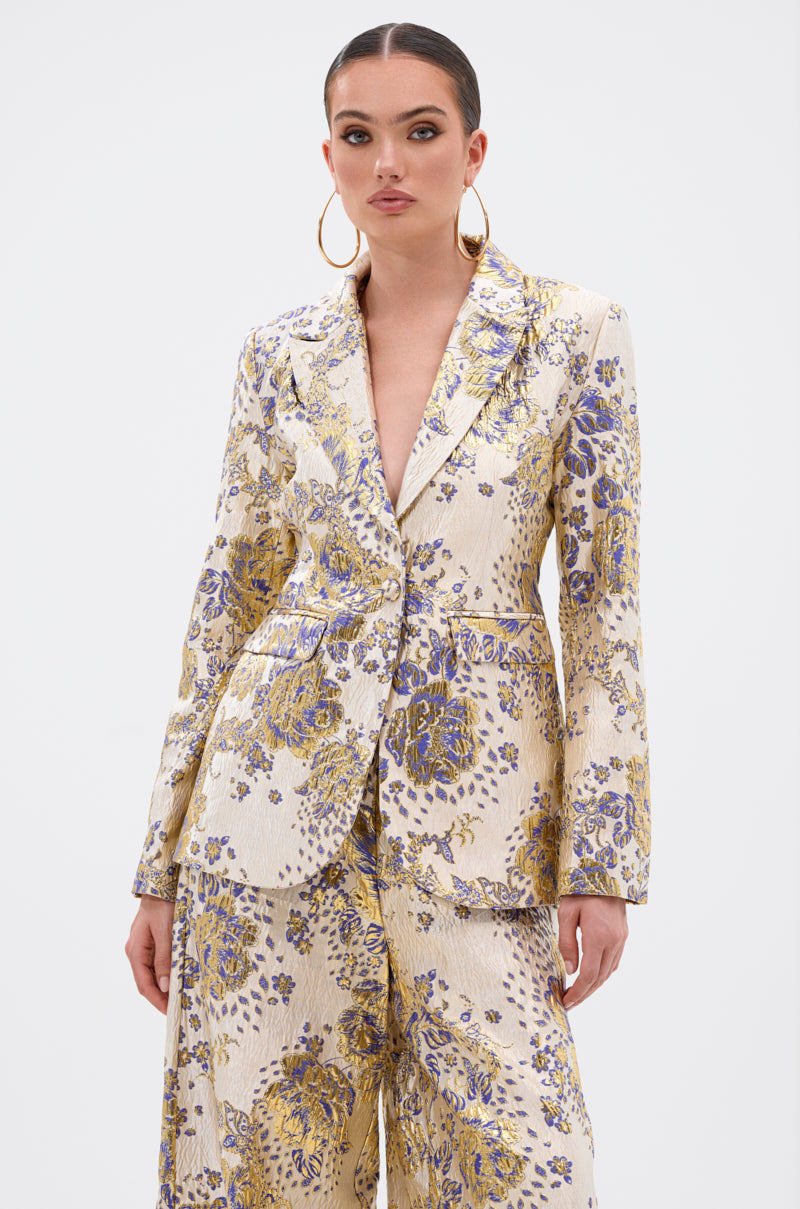 A woman models the GOLDIE BROCADE BLAZER, cream-colored with a purple and yellow floral brocade pattern, paired with matching wide-leg pants. She finishes the look with slicked-back hair, hoop earrings, and metallic loafers.