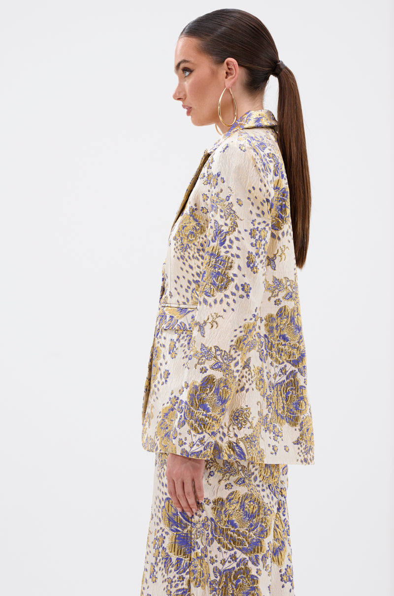 Woman with long brown hair in a low ponytail and large hoop earrings models the GOLDIE BROCADE BLAZER—a cream blazer with purple and gold floral brocade—shown from the side against a plain white background.