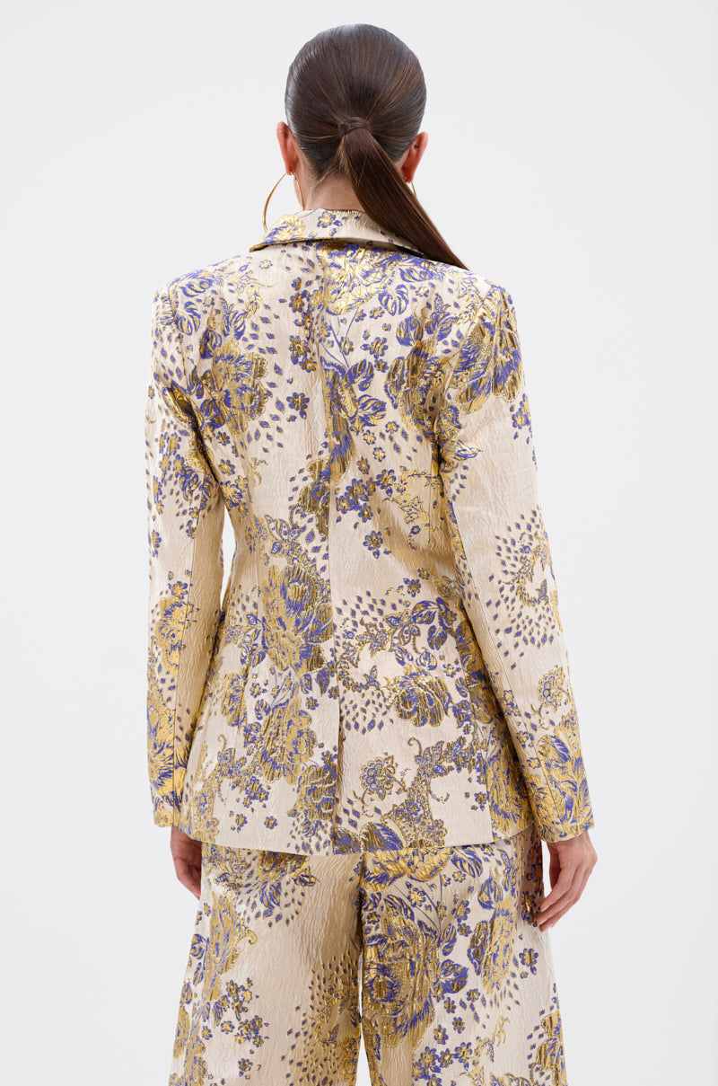 A woman with long dark hair in a low ponytail is seen from behind, wearing the GOLDIE BROCADE BLAZER and matching wide-leg pants, featuring a beige suit with a purple and gold floral brocade pattern for an elegant look.