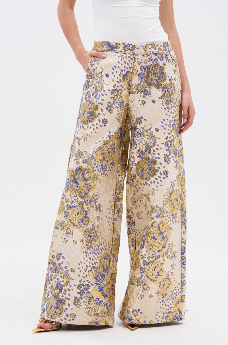 Wearing the GOLDIE BROCADE TROUSER, featuring a beige base with purple and yellow floral brocade, a person stands with one hand in their pocket, paired with a white top and open-toe shoes.