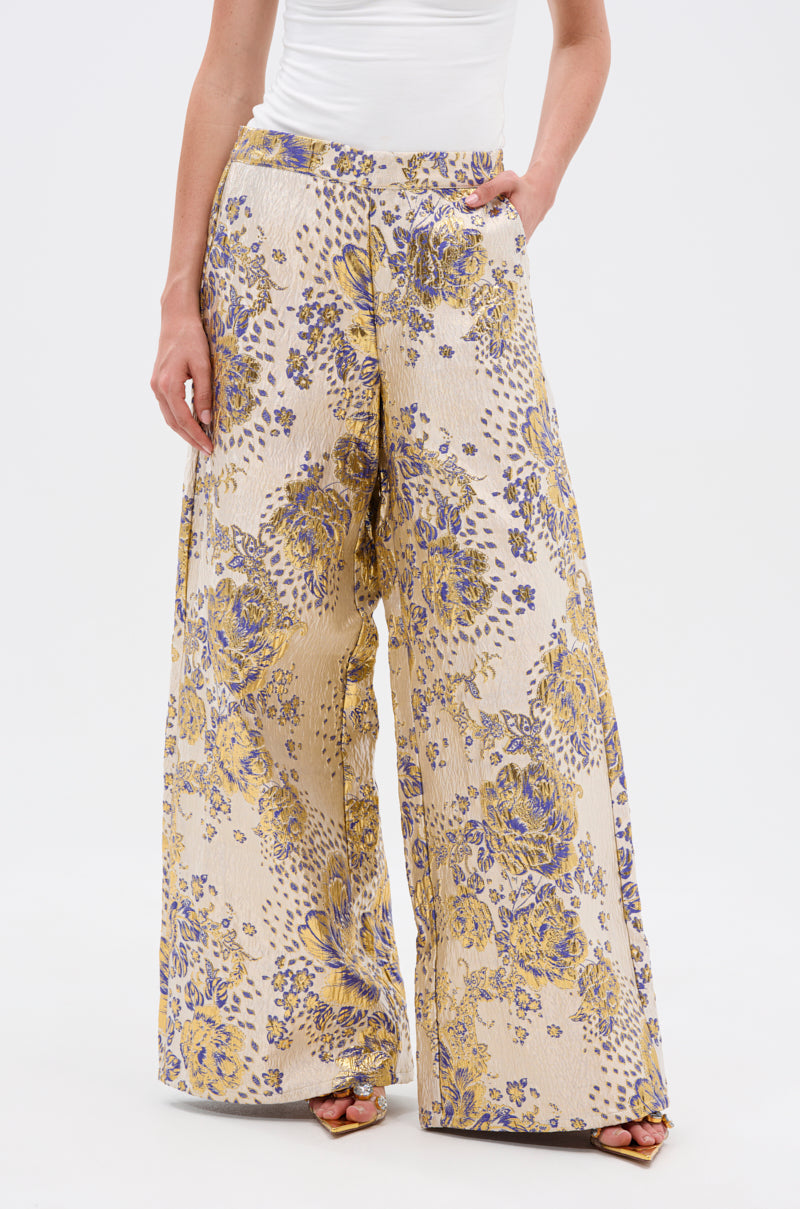 Wearing the GOLDIE BROCADE TROUSER with a wide leg and yellow-purple floral brocade, this person pairs the textured, pocketed pants with a white top, standing against a plain white background.